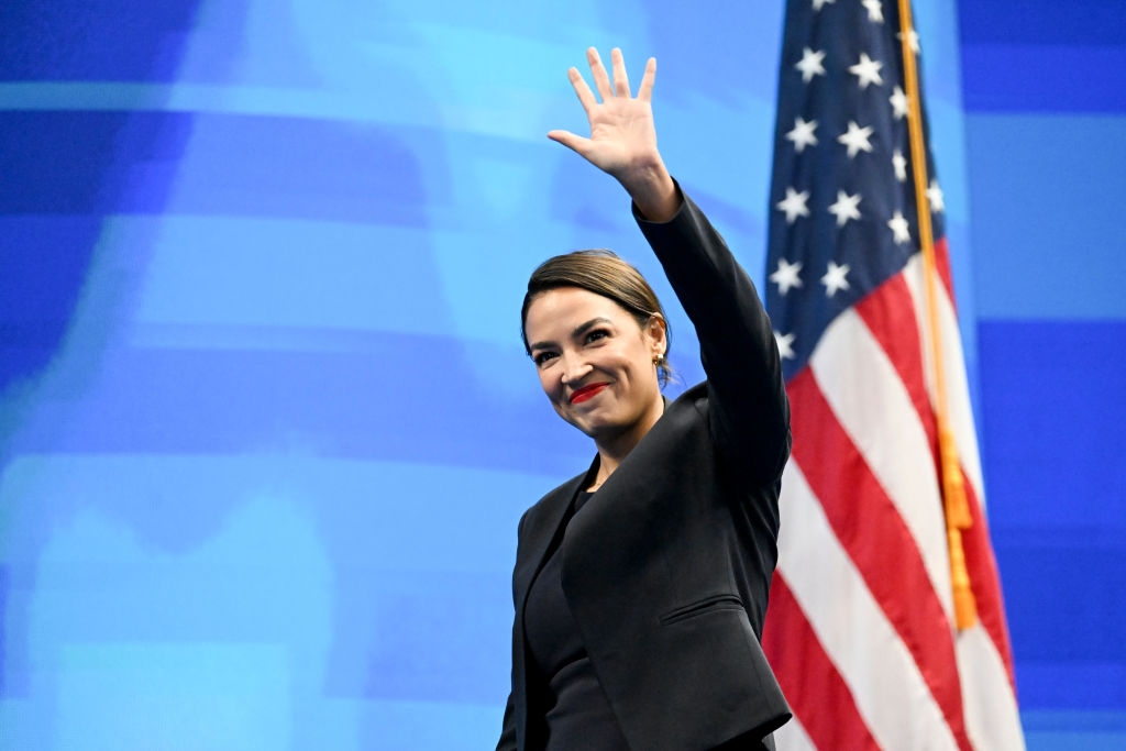 Person in a formal outfit waves on stage with an American flag in the background