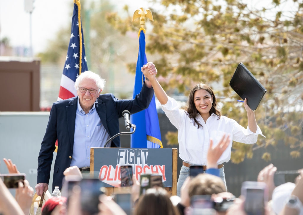 Two people hold hands on stage at a rally. One holds a folder. A crowd watches, and a sign reads "Fight Oligarchy" with flags in the background