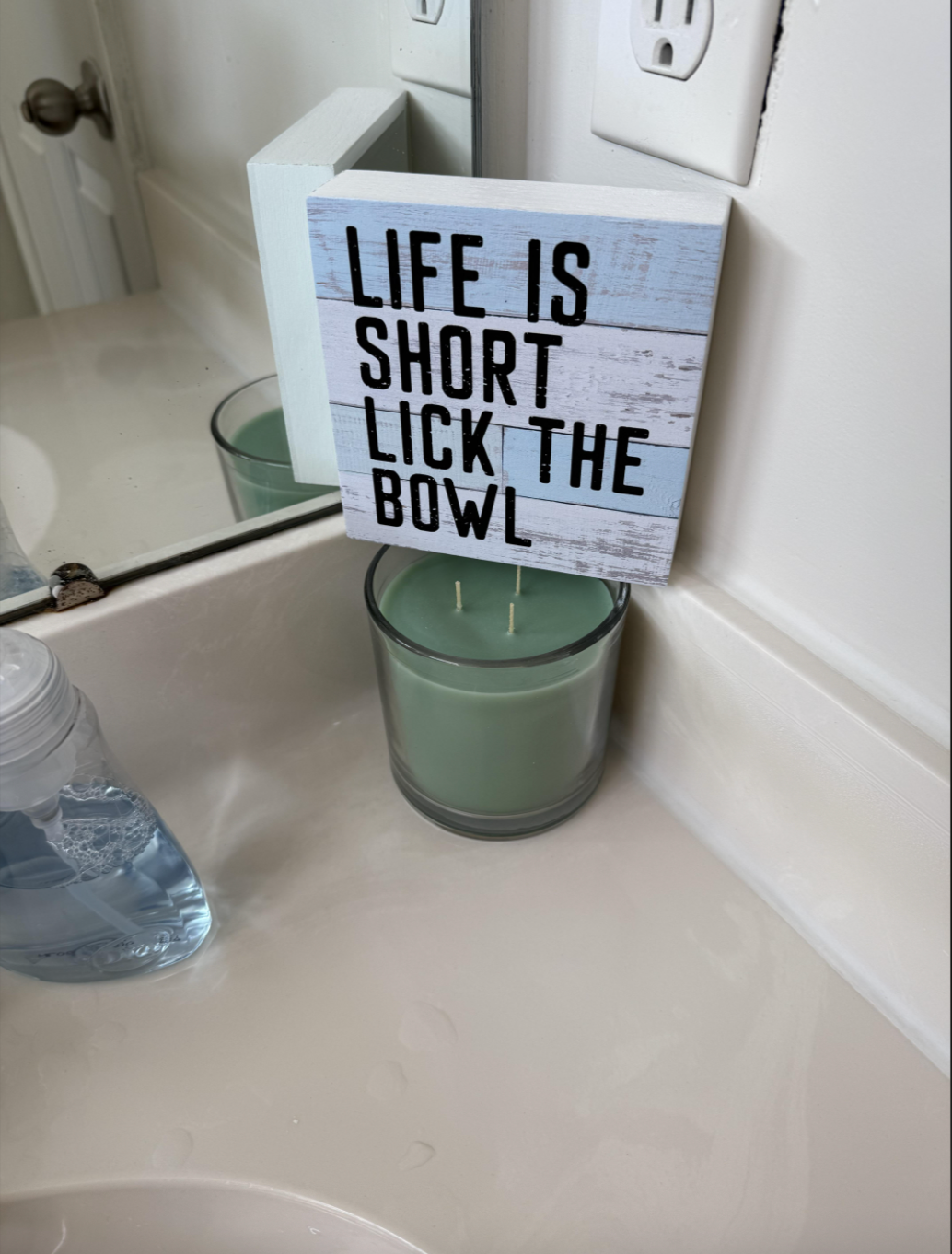 Sign on bathroom counter reads: "Life is short, lick the bowl." It sits above a candle and next to a soap dispenser