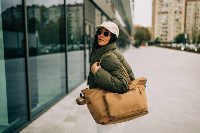 Person wearing a puffy jacket, hat, and sunglasses, carrying a large bag, walking by a modern building