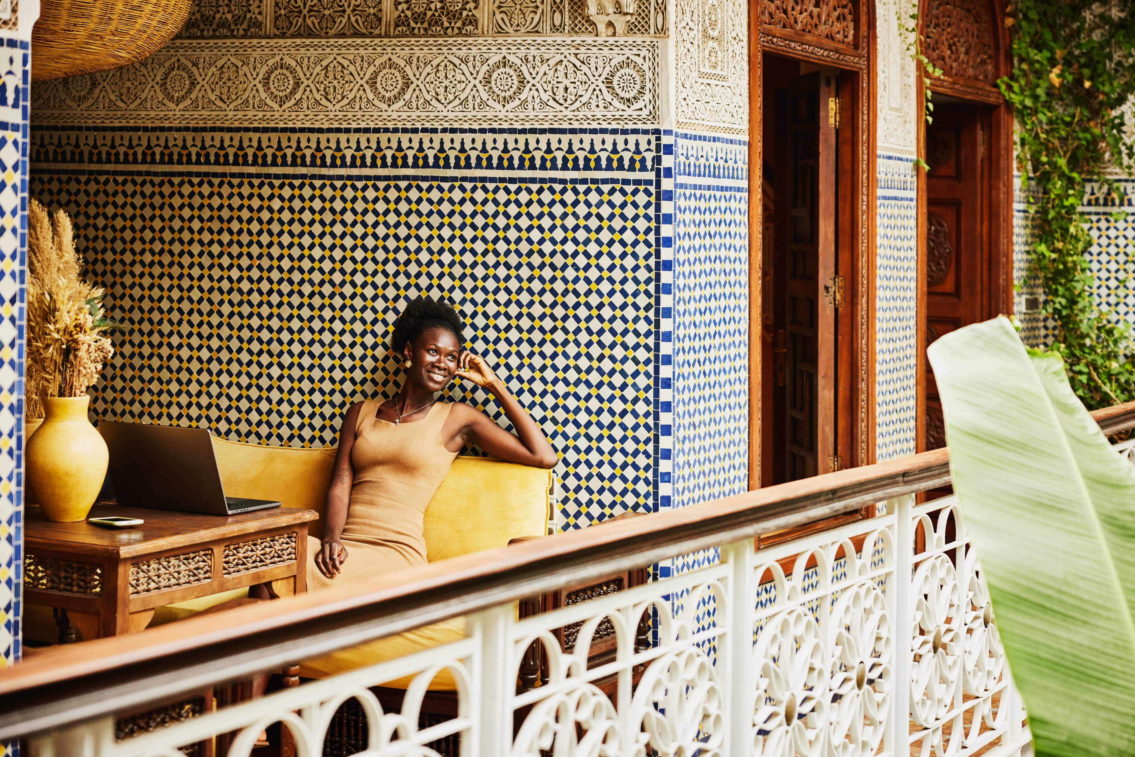 Person sitting on a balcony with intricate patterned walls, smiling and relaxed. A laptop is on the table beside them