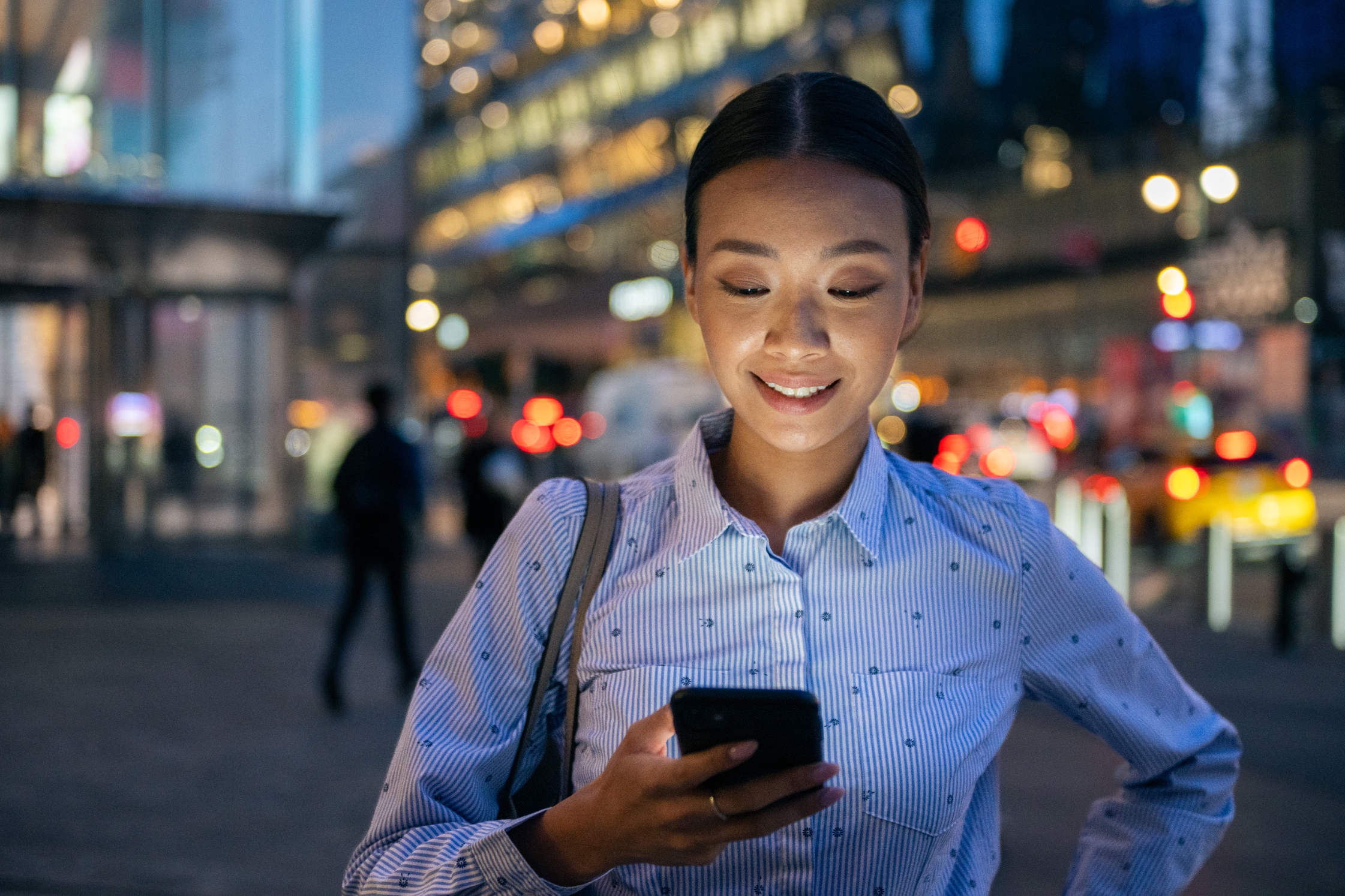 Person smiling while looking at a smartphone on a city sidewalk at night, with blurred traffic and lights in the background