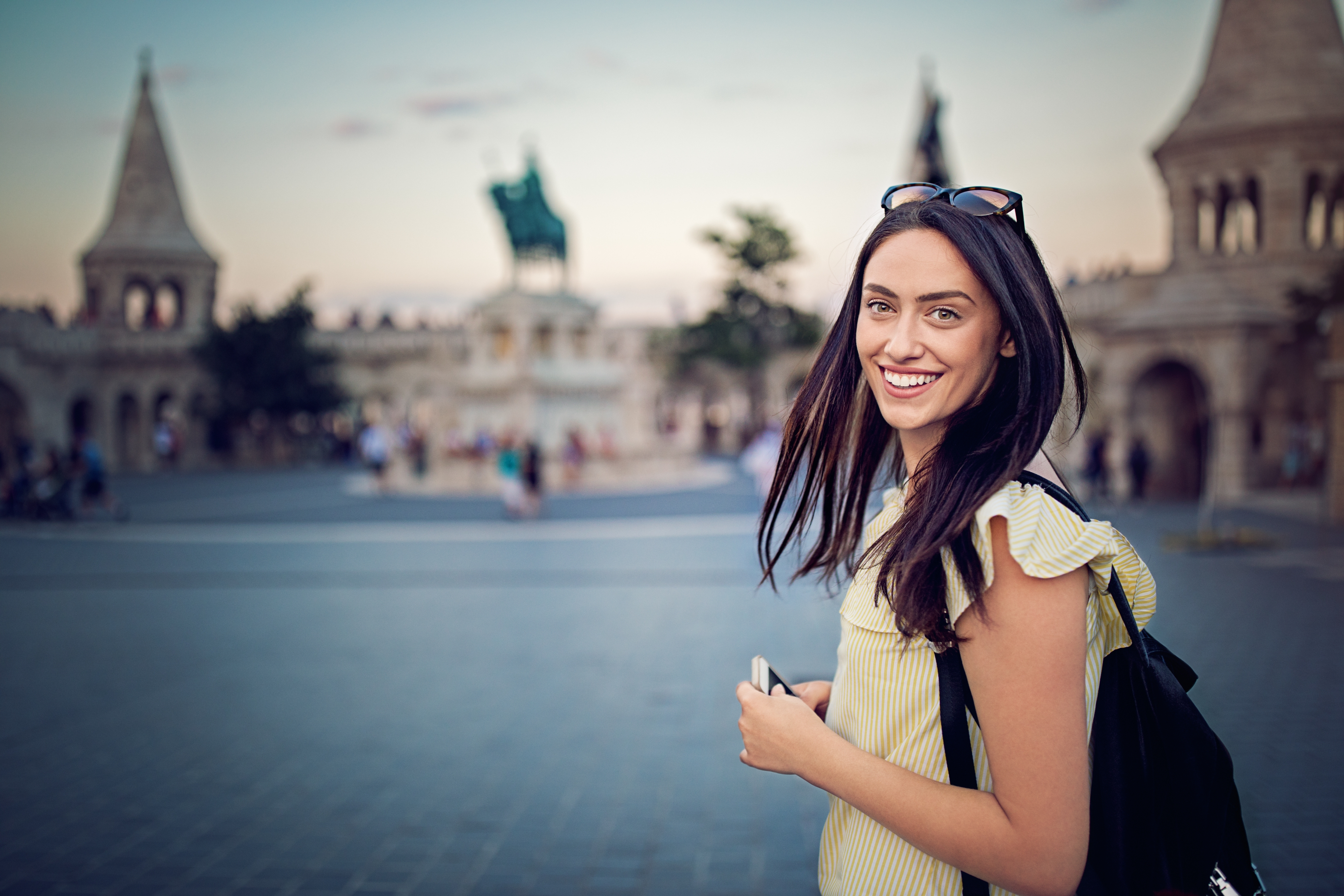 Person smiling outdoors at a historic site with pointed towers, holding a phone and wearing a casual summer outfit