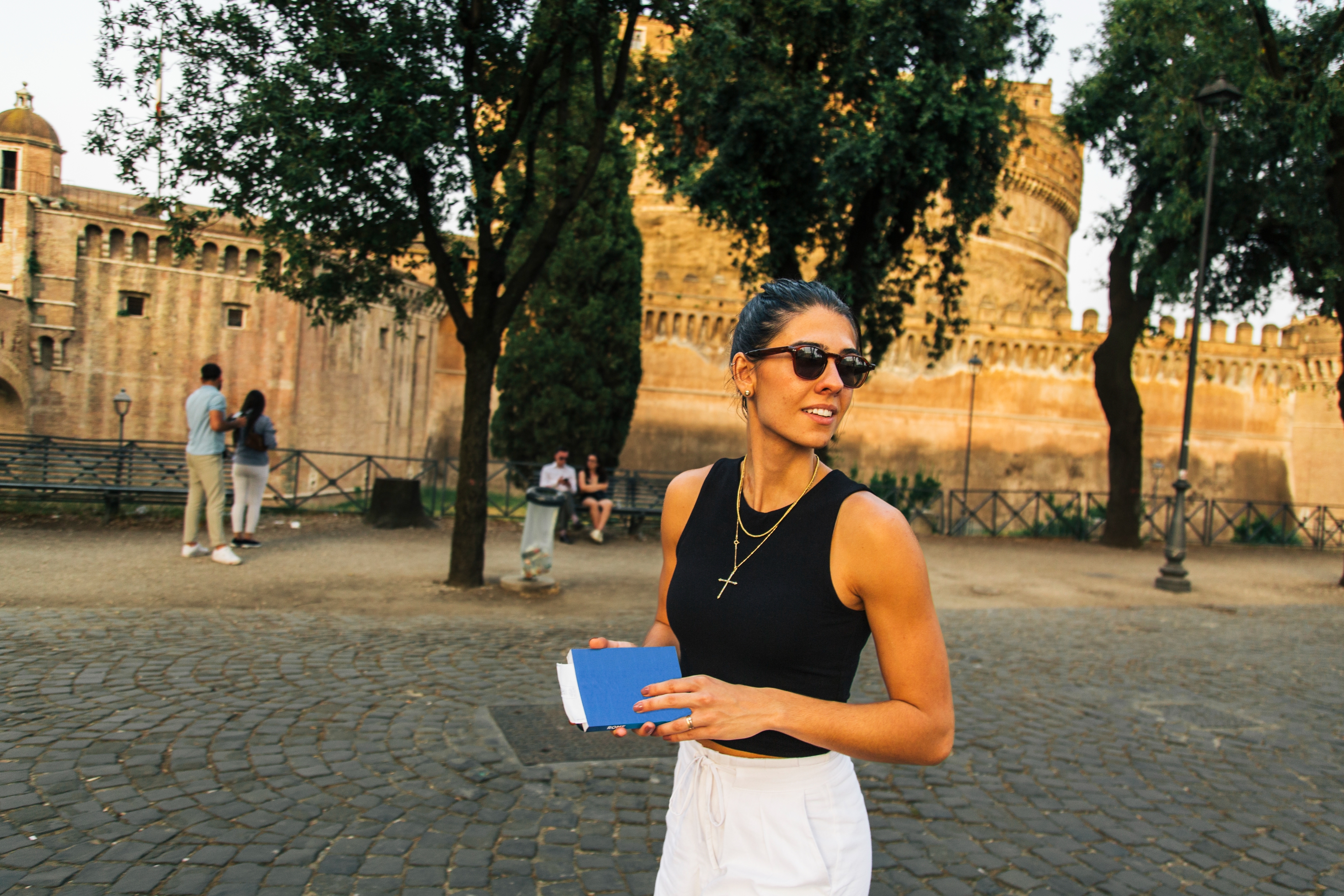 A woman in sunglasses, wearing a sleeveless top and white pants, holds a book in front of a historic castle backdrop