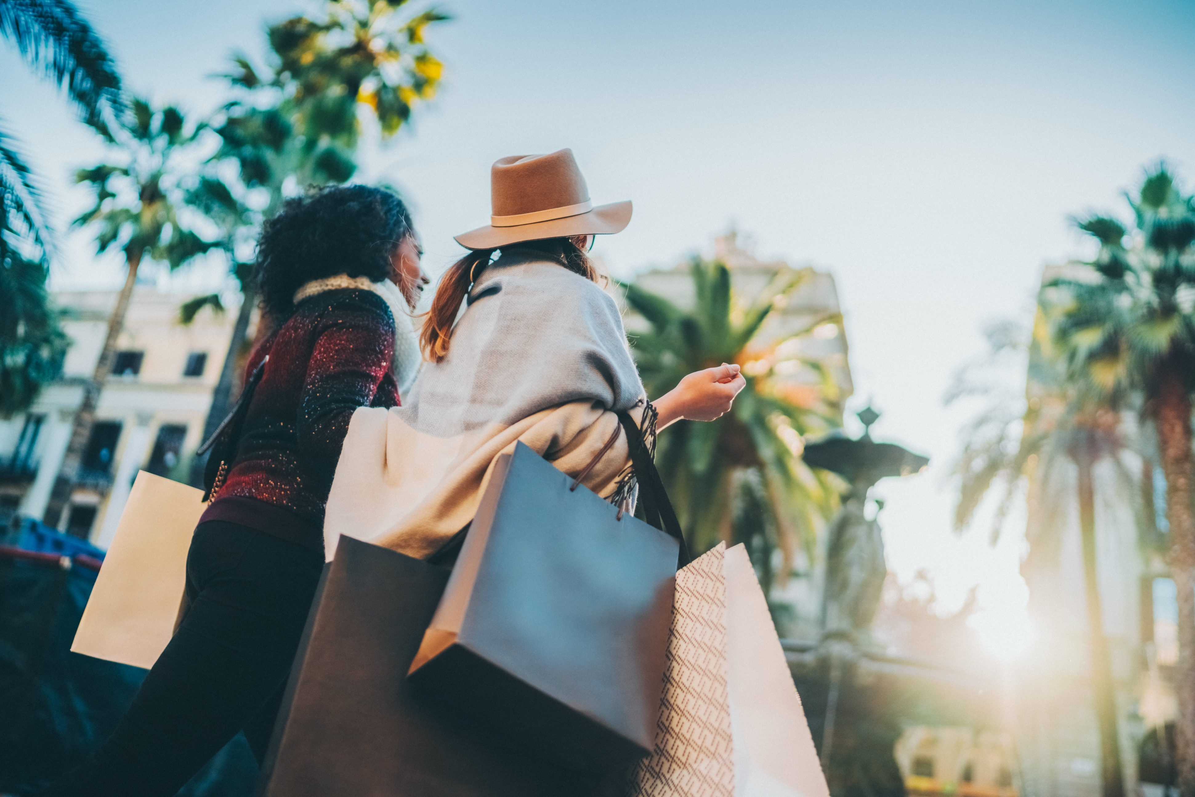 Two people with shopping bags stroll through a sunny outdoor plaza with palm trees. One wears a hat and a shawl