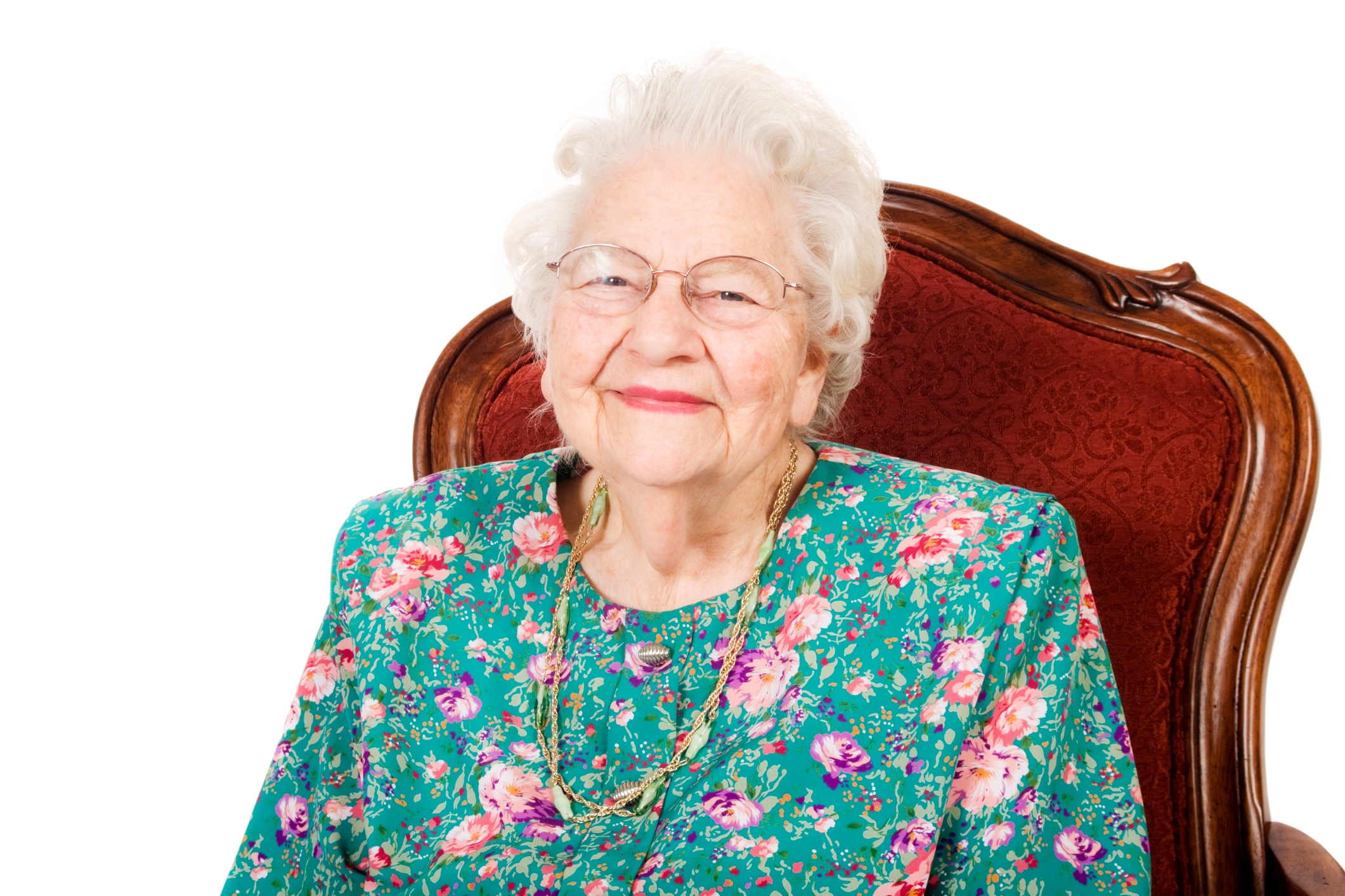Elderly woman with glasses, smiling, sitting in a chair, wearing a floral-patterned top and necklace