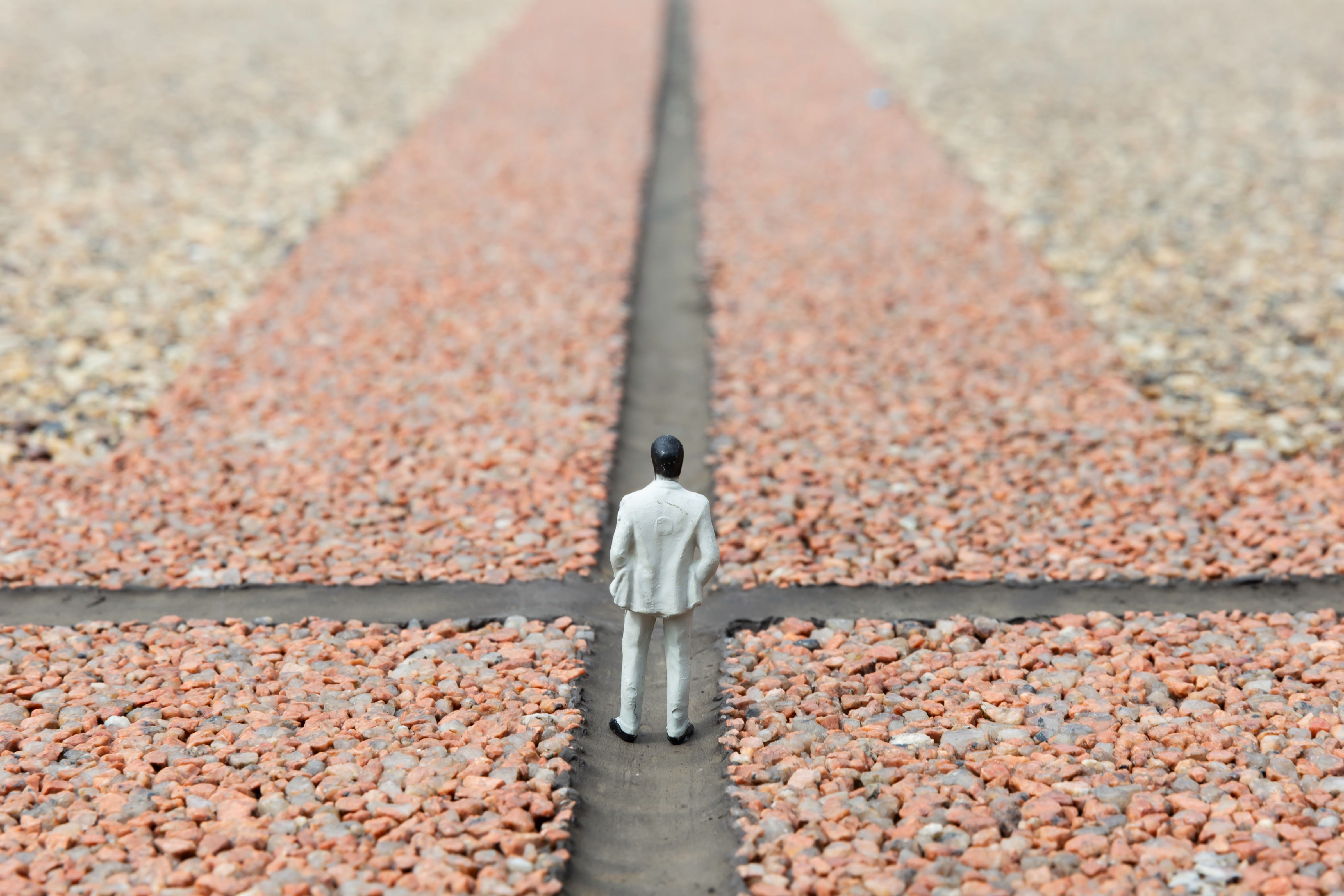 A miniature figure of a person in a suit stands at a crossroads on a gravel path, symbolizing decision-making or choices