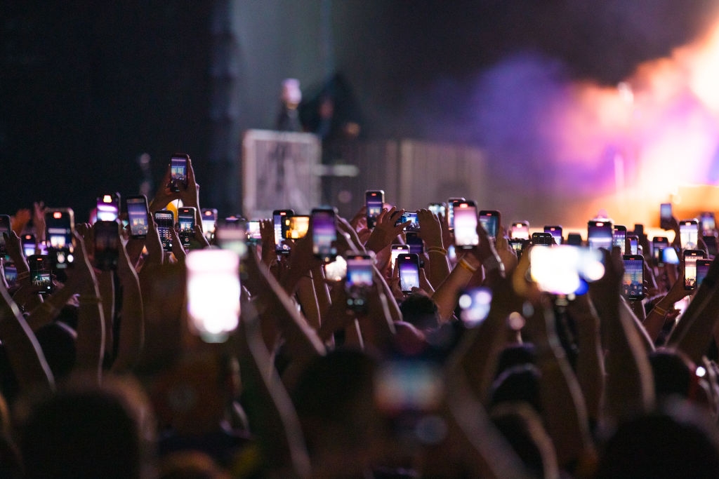 Audience members hold phones up, capturing a live concert performance. The stage is partially visible amidst a sea of raised arms and glowing screens
