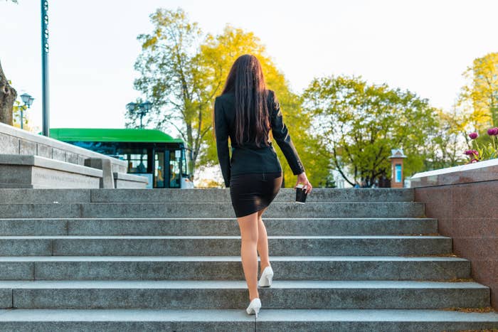 Person in a blazer and skirt walking up outdoor stairs, holding a small clutch, with trees and a building in the background
