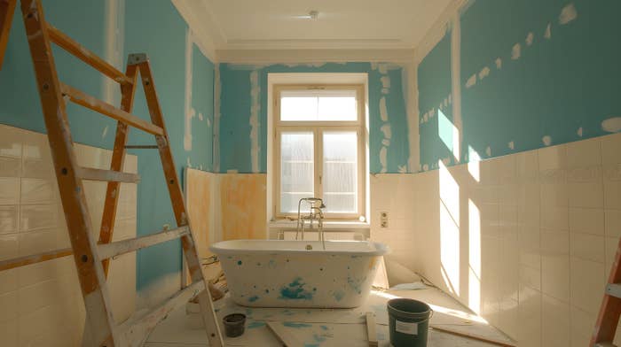 Bathroom under renovation with ladder, bathtub, unfinished walls, and supplies scattered around. Sunlight streams through a window, highlighting the changes
