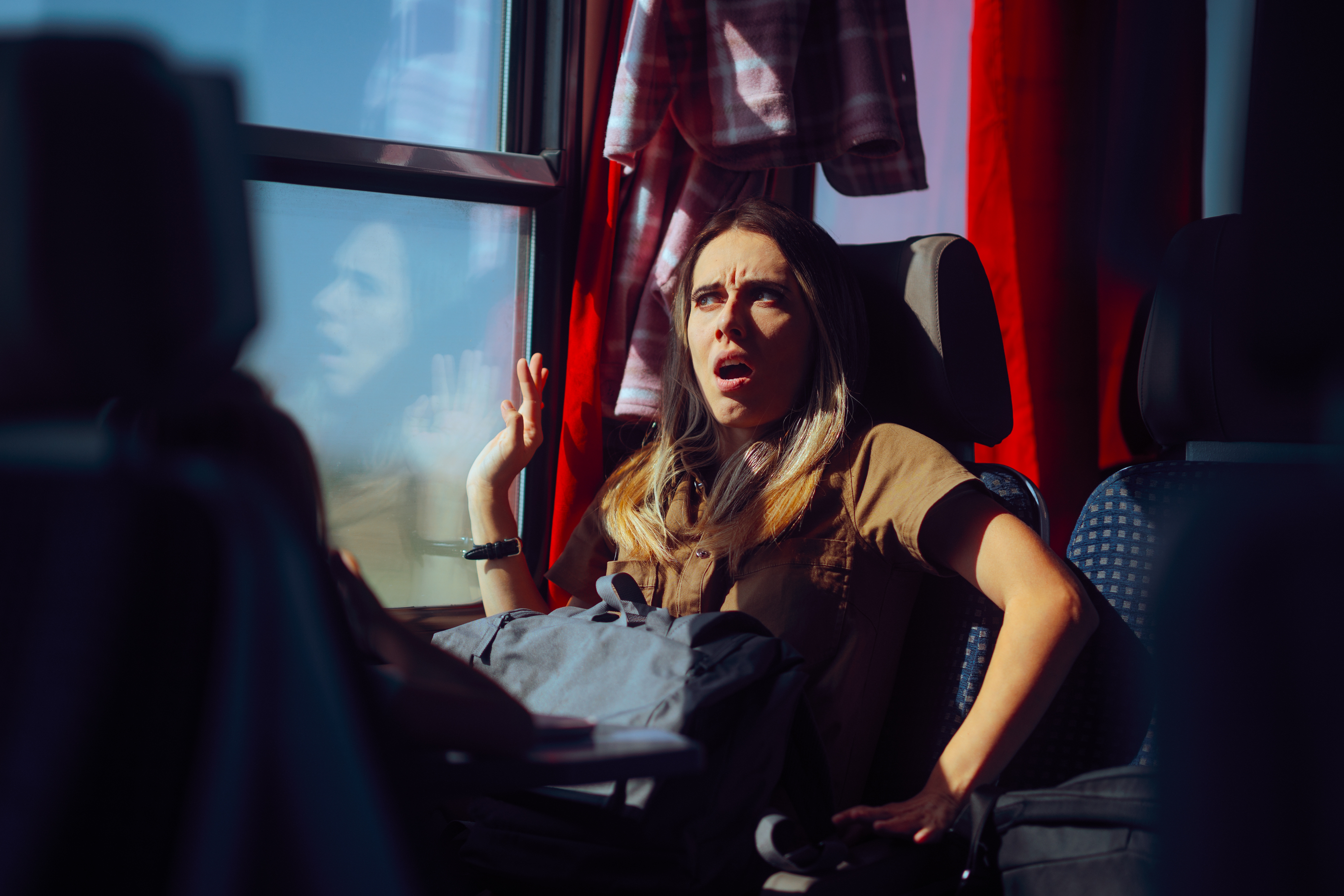 Person on a train looks surprised, sitting by the window. Sunlight streams in, casting shadows. A plaid shirt hangs in the background
