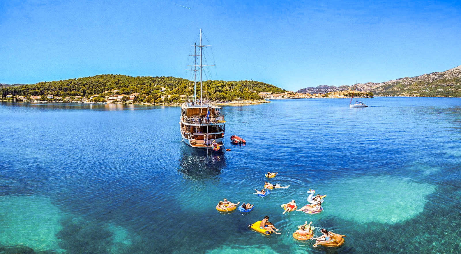 People enjoy water activities near a large boat in a scenic, calm ocean setting with lush green hills in the background