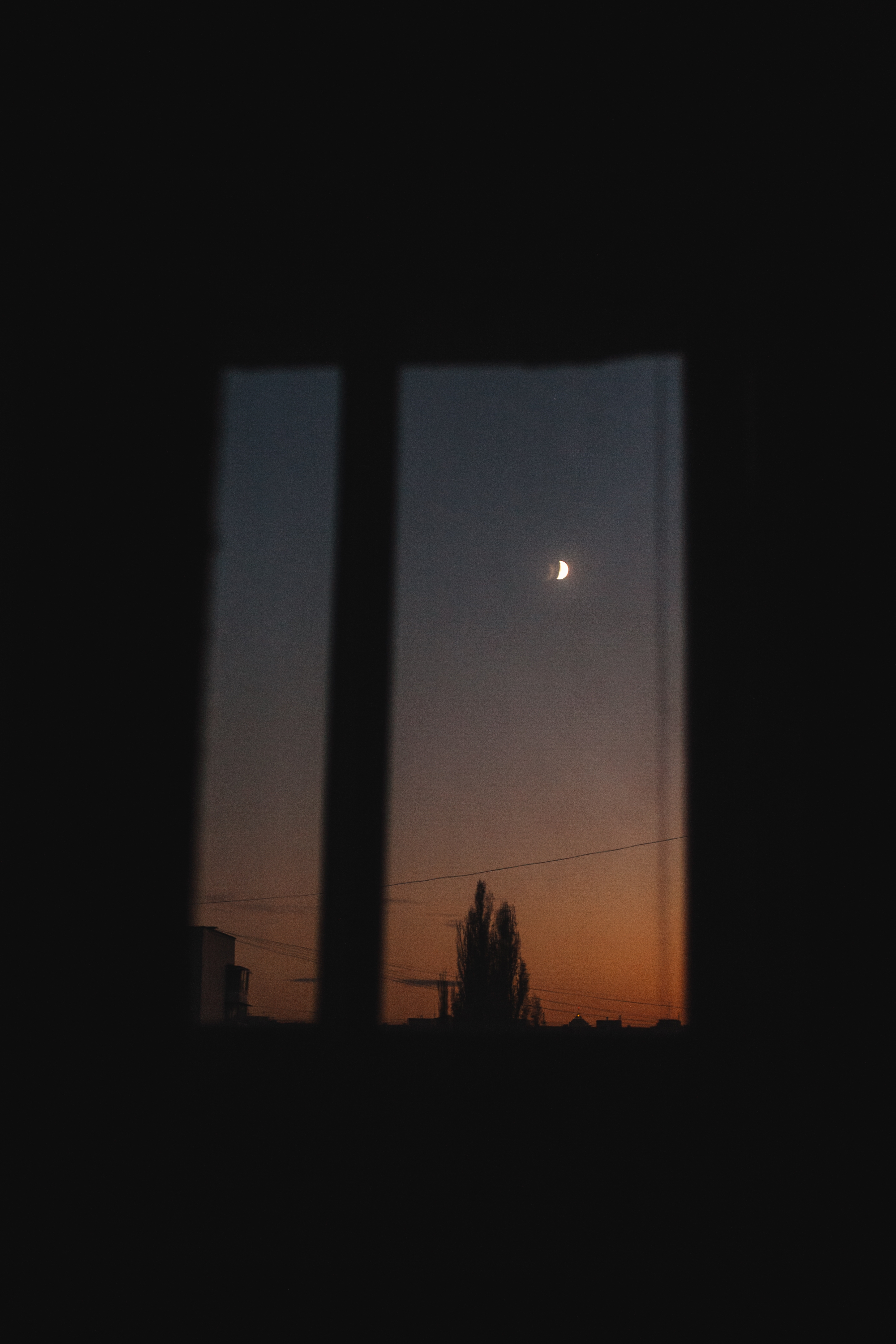 Crescent moon visible through a window at dusk, with a silhouette of trees and buildings in the background