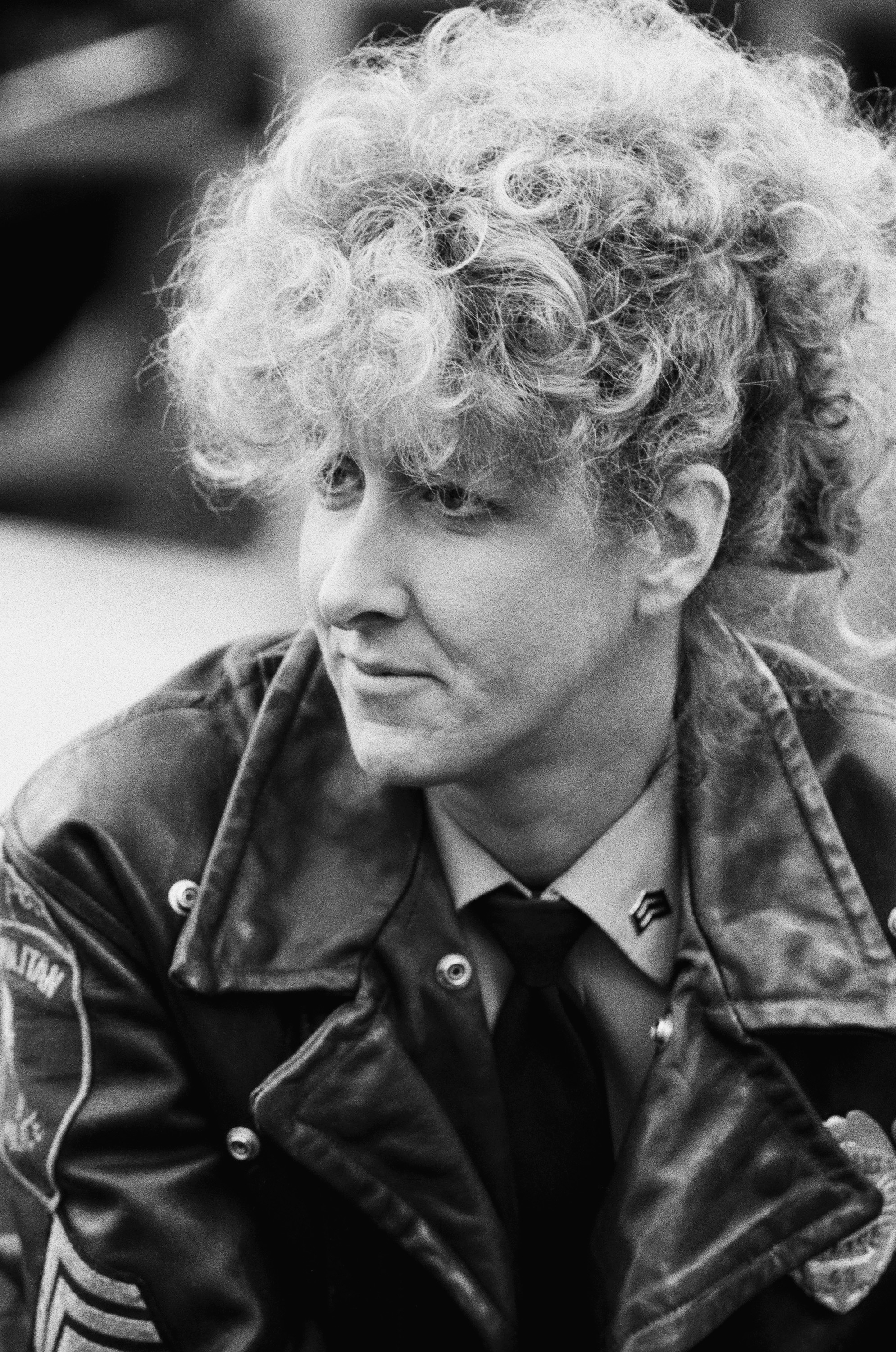 Person with curly hair in a leather jacket, possibly a law enforcement uniform, looking thoughtfully to the side