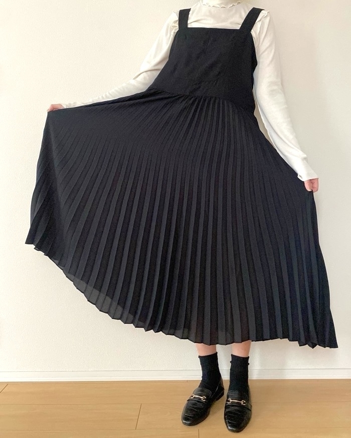 白い長袖シャツに黒のプリーツジャンパースカートを着た人物が、スカートの裾を持ってポーズを取っている。