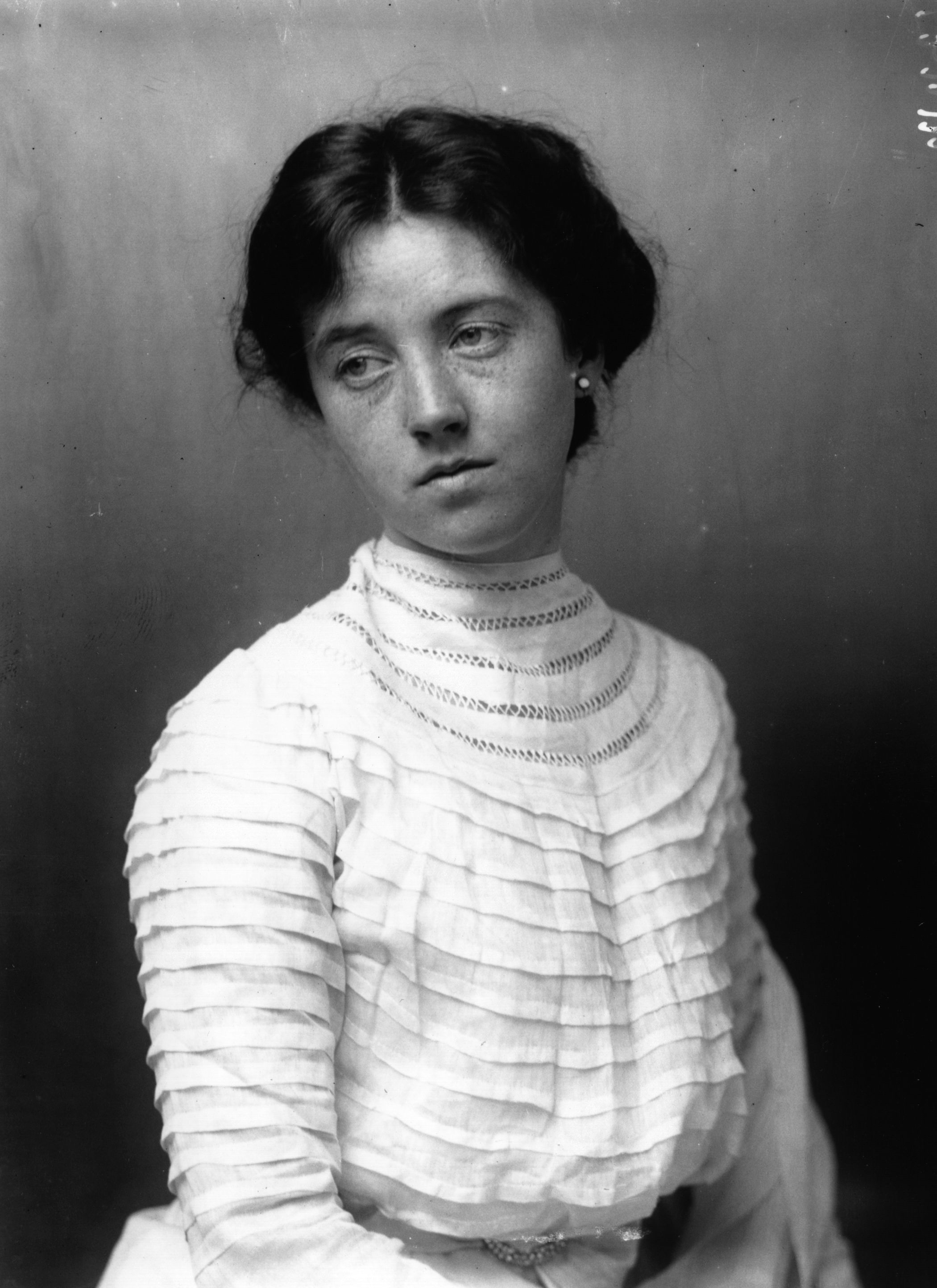 A woman in early 1900s attire with a high-necked, pleated blouse gazes off camera, with a neutral expression