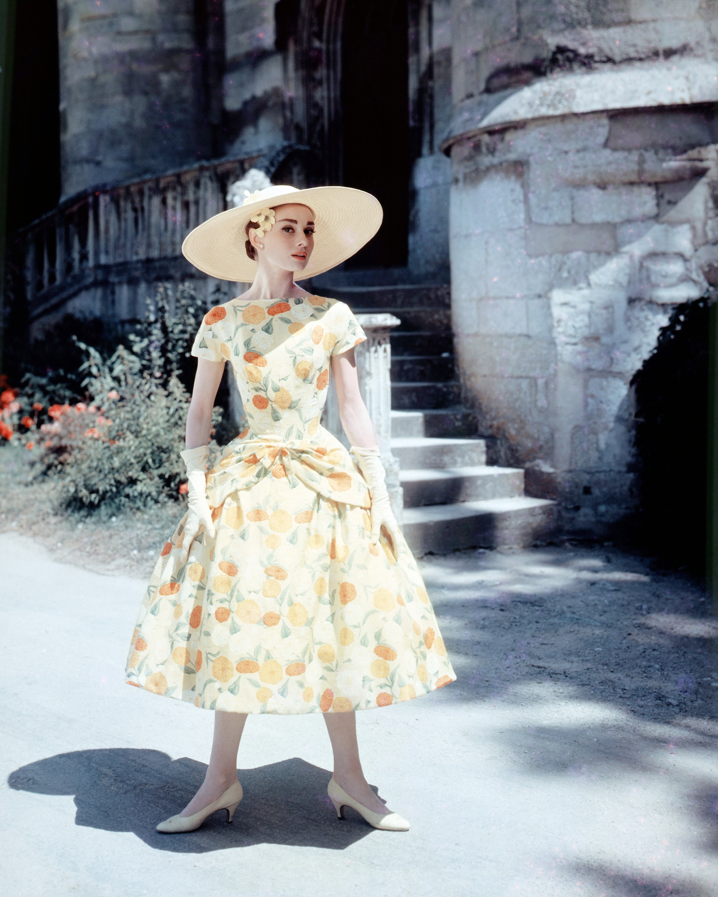 Person in vintage floral dress and wide-brimmed hat standing outdoors near stone steps, exuding elegance and classic style