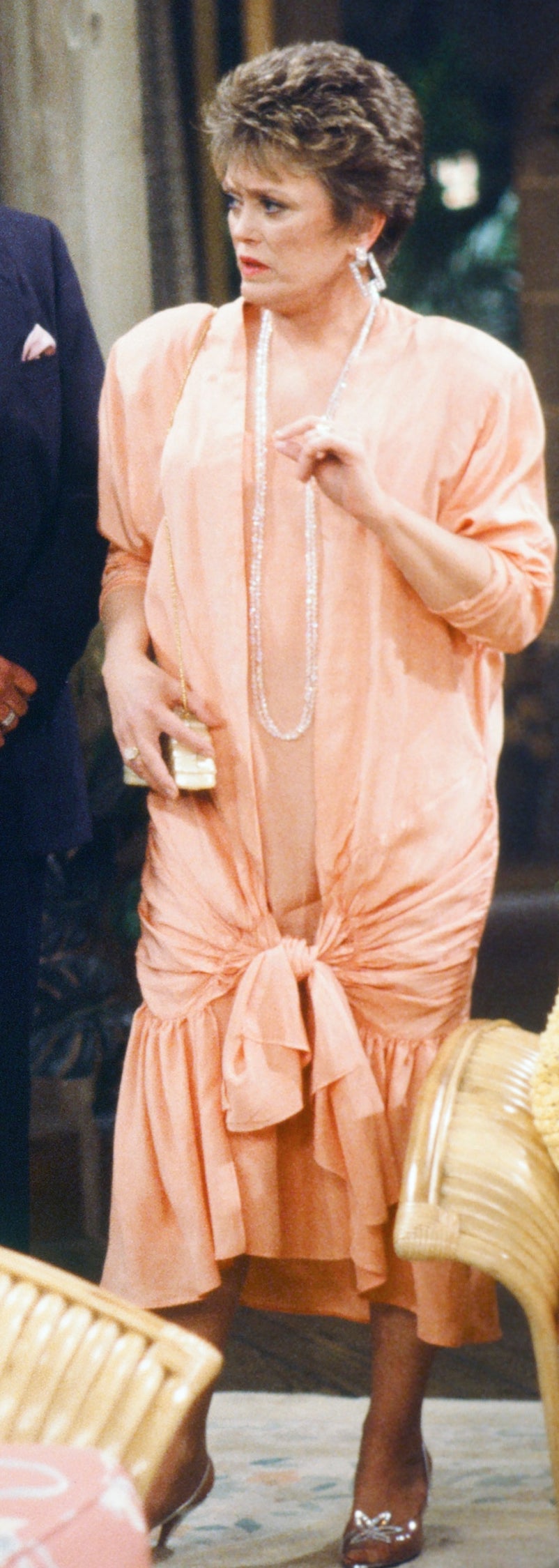 Three people stand in a living room; two men in suits and a woman in a ruched dress holding an item, engaged in conversation