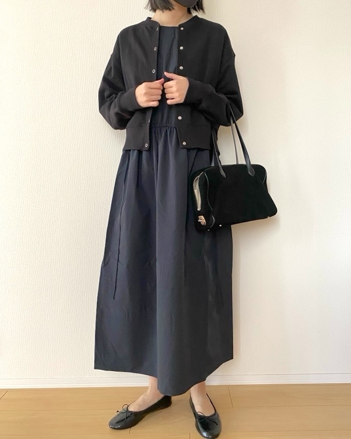 女性がシンプルなドレスにカジュアルなジャケットを着用し、バッグを持って立っている。ドレスと靴は落ち着いたデザイン。