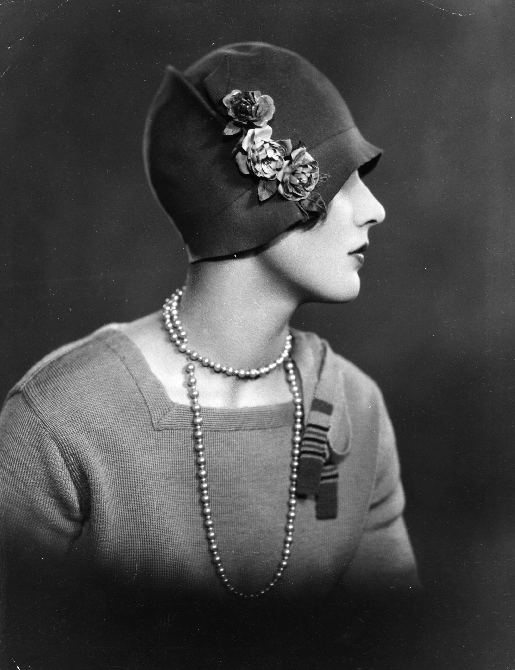 Person in a vintage photo wearing a cloche hat adorned with flowers, a pearl necklace draped, facing sideways. Their attire is formal and elegant