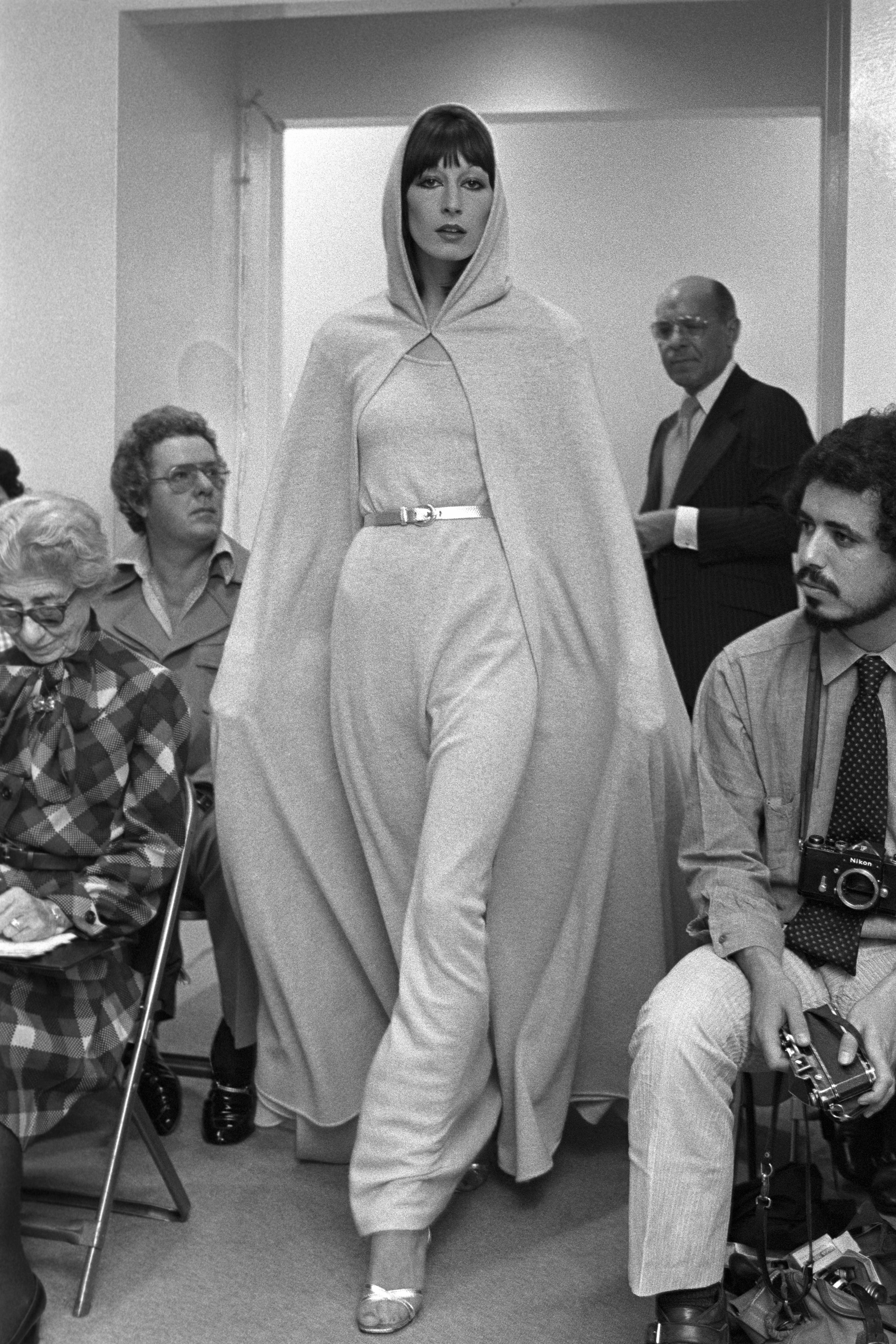 A model walks in a fashion show wearing a hooded cape and belted jumpsuit. People seated nearby, some with cameras, observe intently