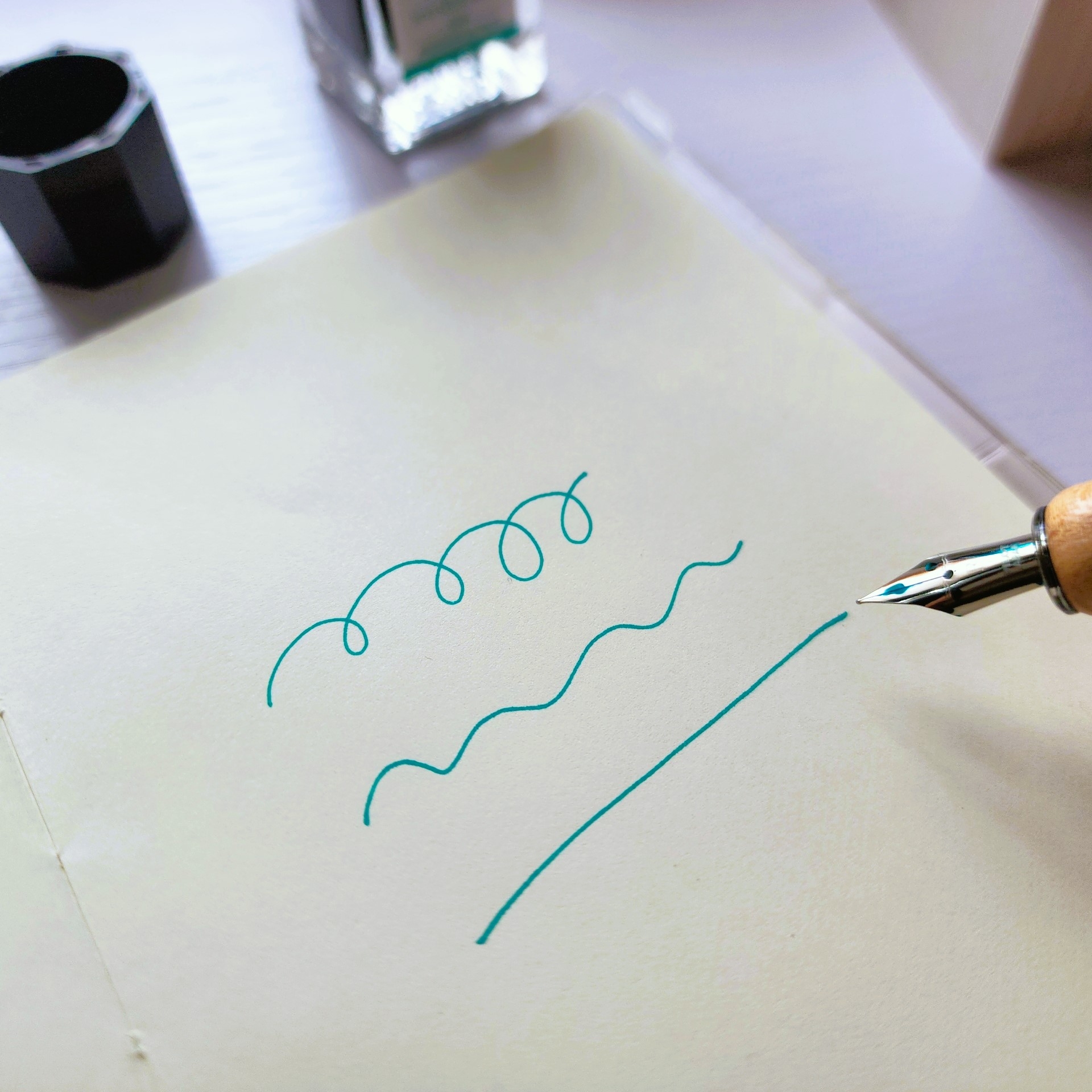 ペンがノートに波線と渦巻きを描いている様子。インク瓶が背景に見える。