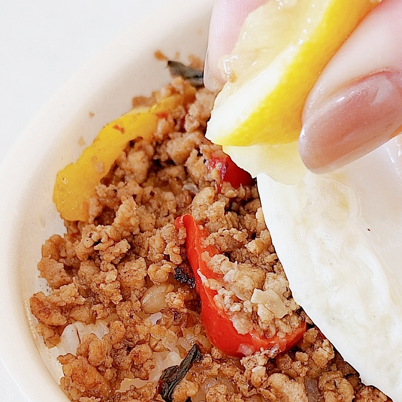 料理の上にレモンを絞る手。挽肉と赤と黄の野菜が見える。目玉焼きも添えられている。