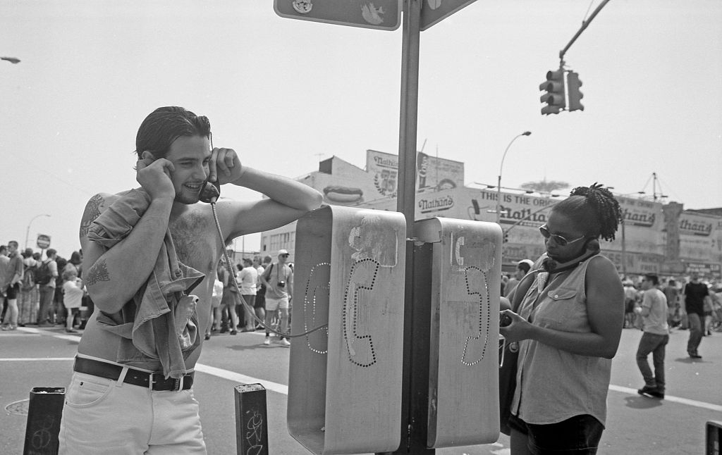 A vintage scene shows a man using a payphone and a person nearby on a cell phone. A busy street and Coney Island's Nathan's Famous are visible