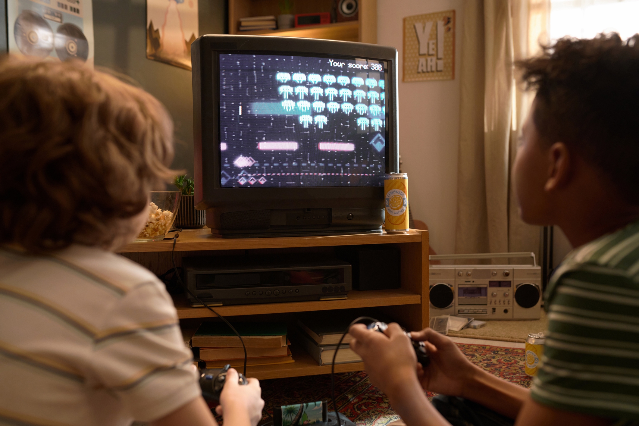 Two people playing a vintage arcade game on a CRT TV, using controllers. A soda and a boombox are nearby, suggesting a nostalgic setting