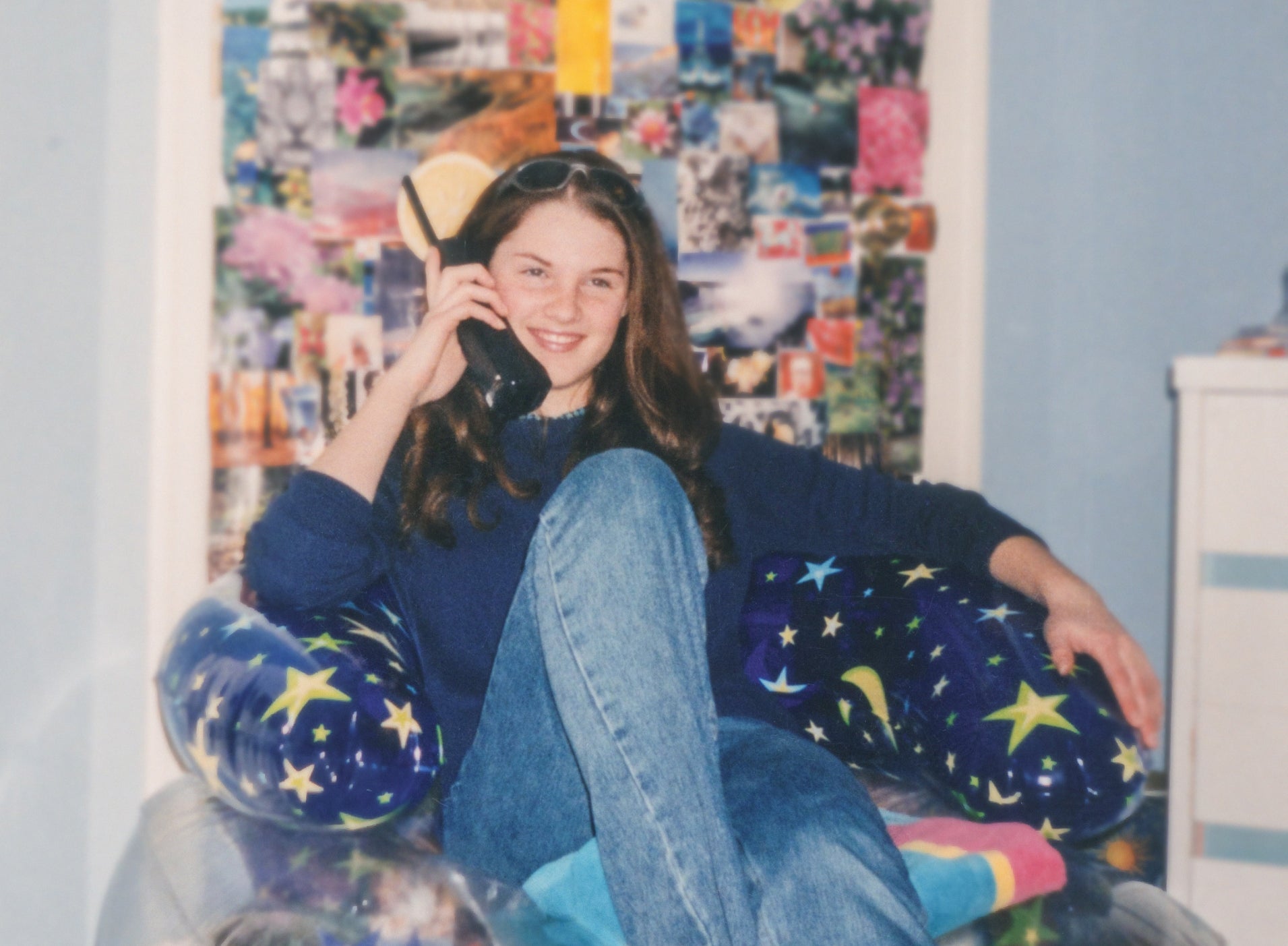 A person sits on a transparent inflatable chair, talking on the phone, with a collage of photos on the wall behind them
