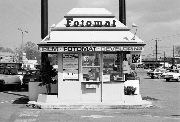 Fotomat kiosk in a parking lot, offering film developing services with various signs promoting film and flash sales