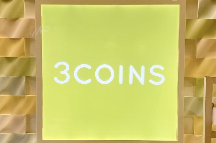 「3COINS」のロゴが中央にある看板。背景には緑色のパネルが並ぶ。