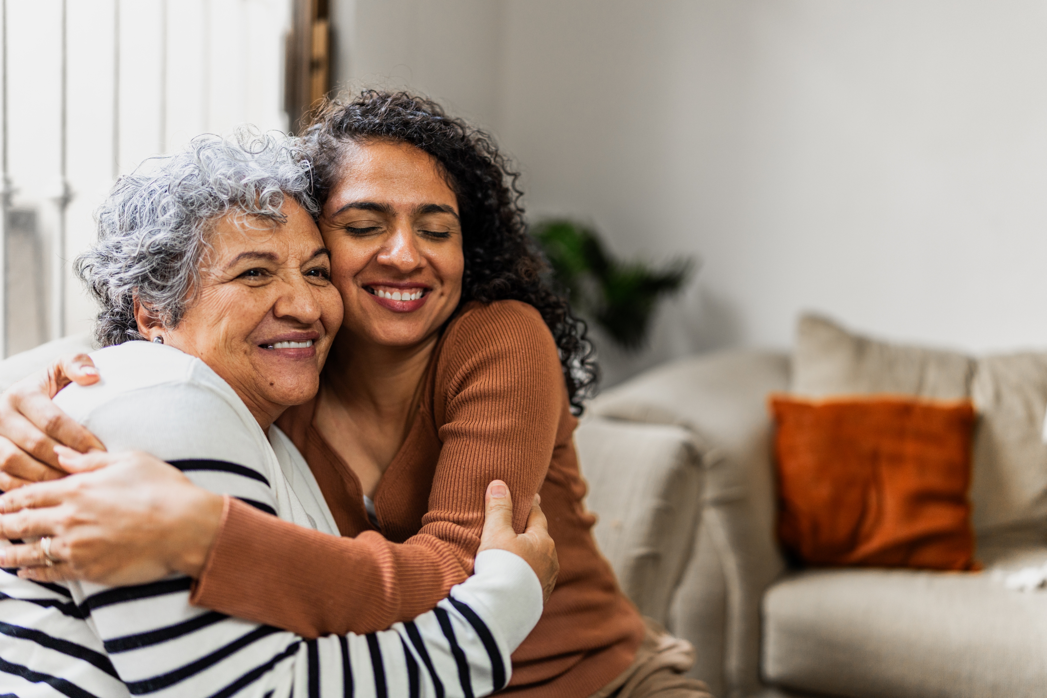 Two women smiling and hugging warmly on a sofa in a cozy living room setting
