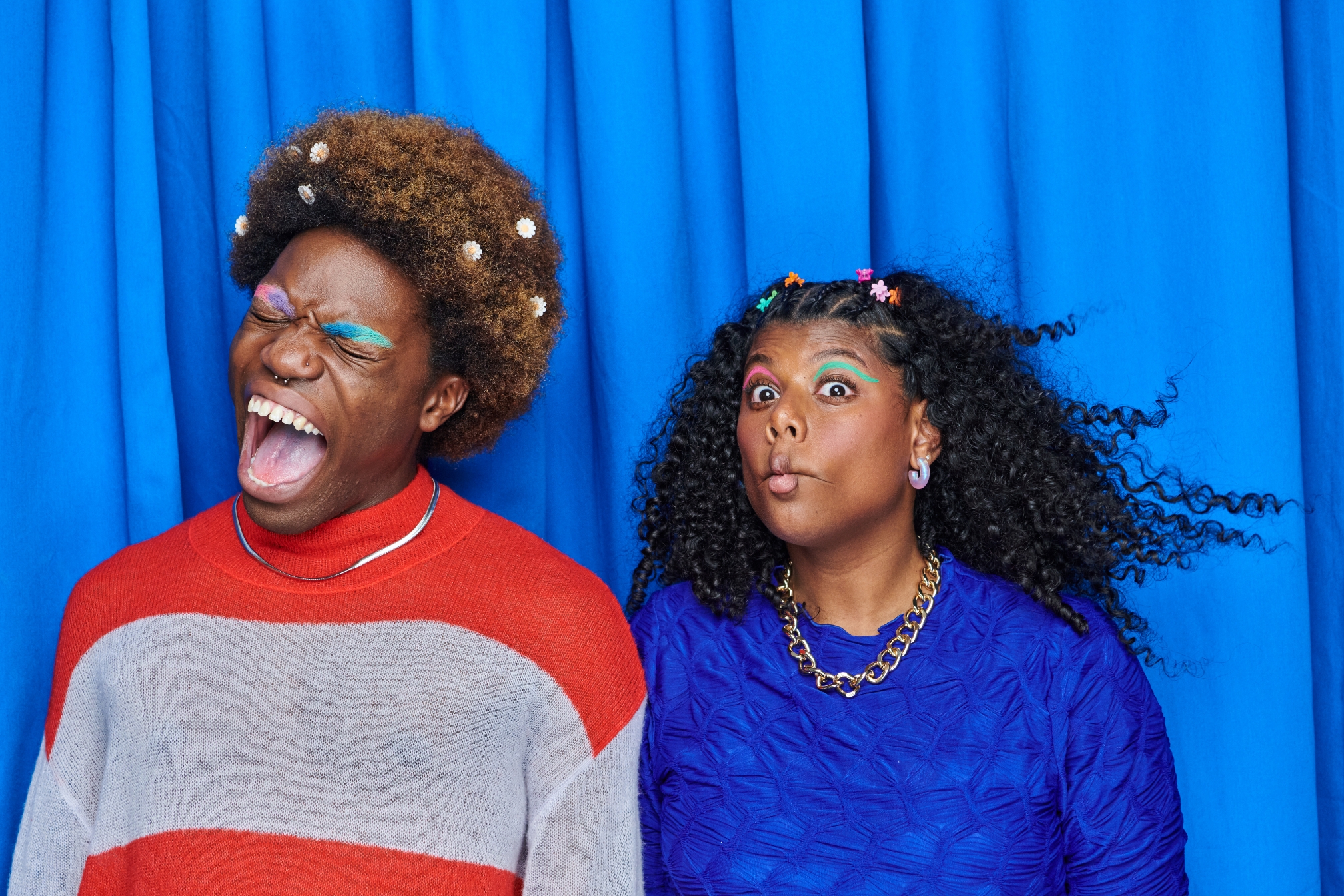 Two people pose against a plain backdrop. One wears a striped sweater and shouts, while the other in a patterned top looks playfully surprised