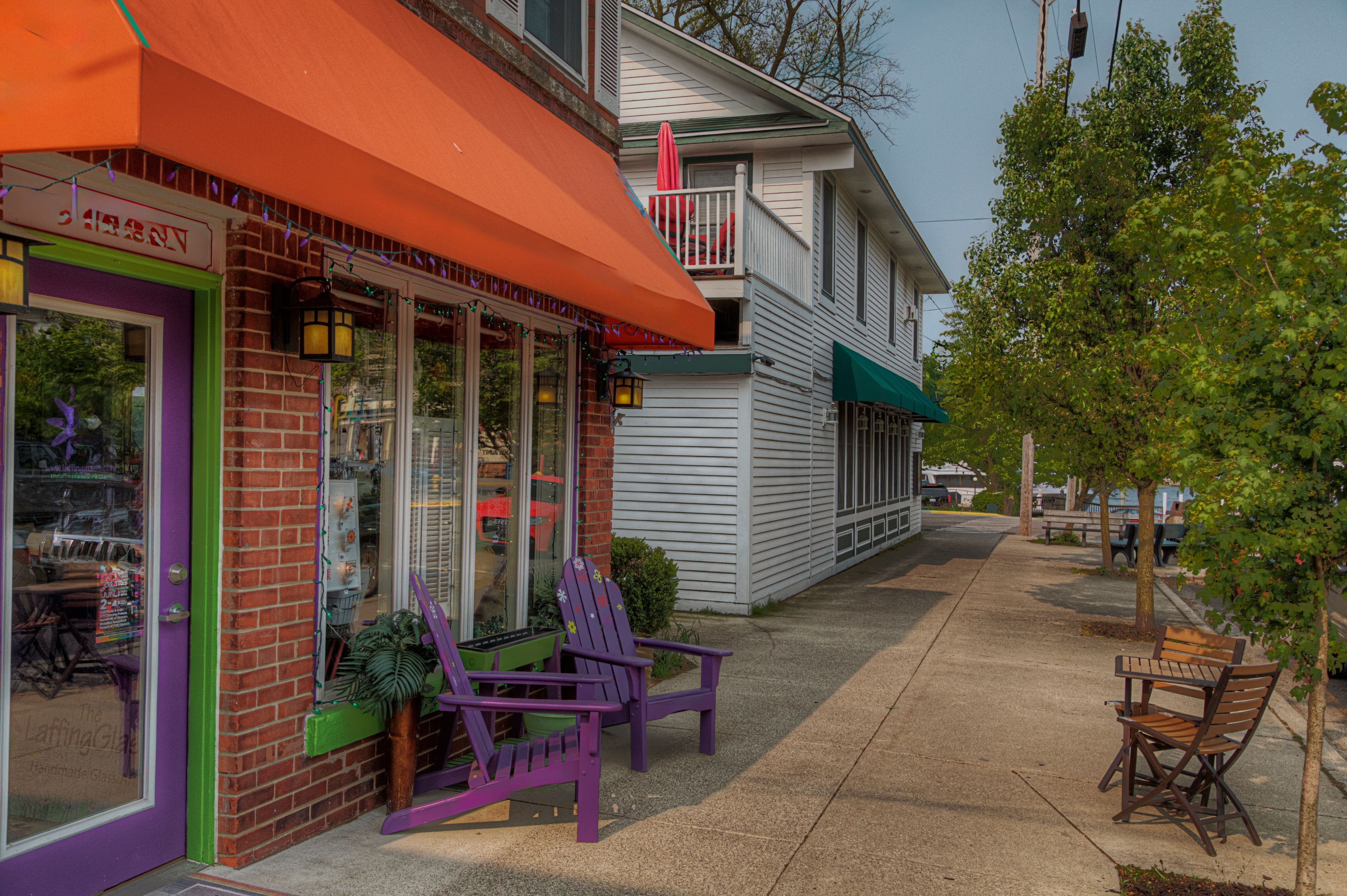 Street view with a sidewalk café, featuring outdoor seating and storefronts. Trees line the path, contributing to a quaint, inviting atmosphere