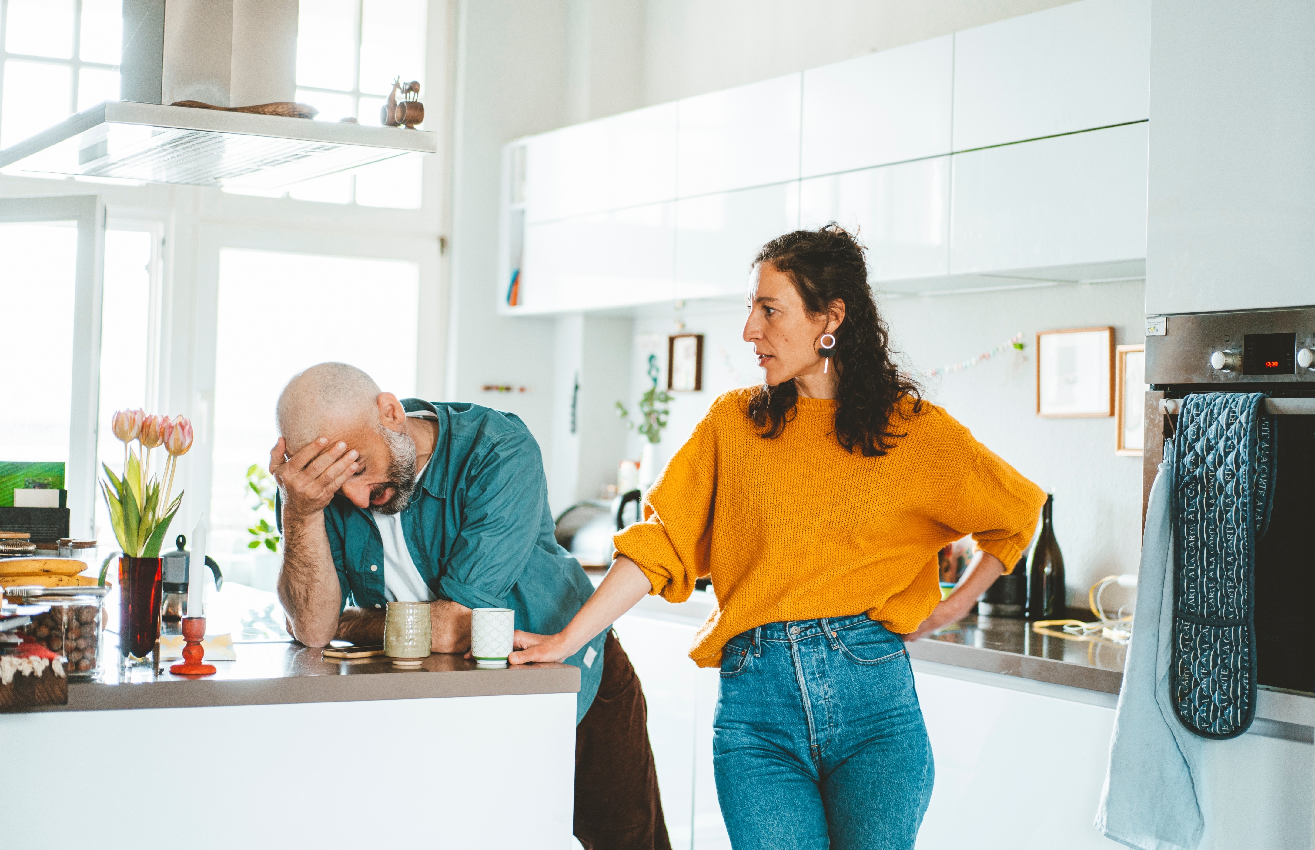 A woman in a casual sweater and jeans talks to a tired-looking man leaning on a kitchen counter, suggesting a serious conversation