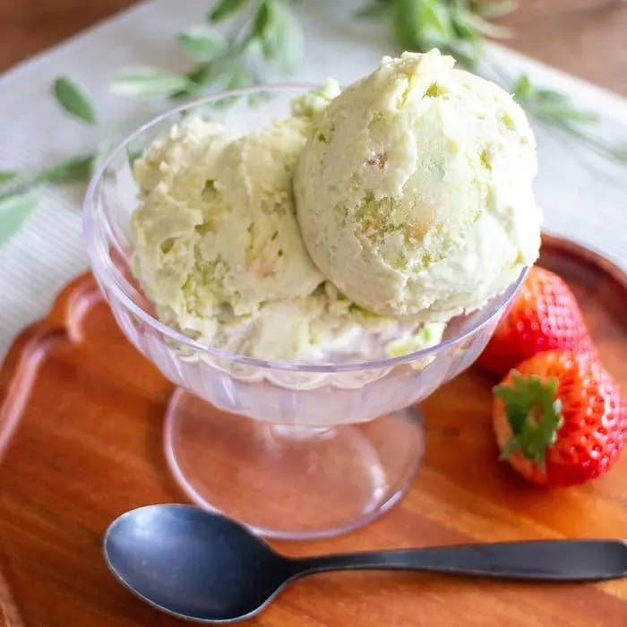 ガラスの器に盛られたピスタチオアイスクリームの隣にイチゴが添えられ、木製のトレイに置かれたスプーンが見えます。