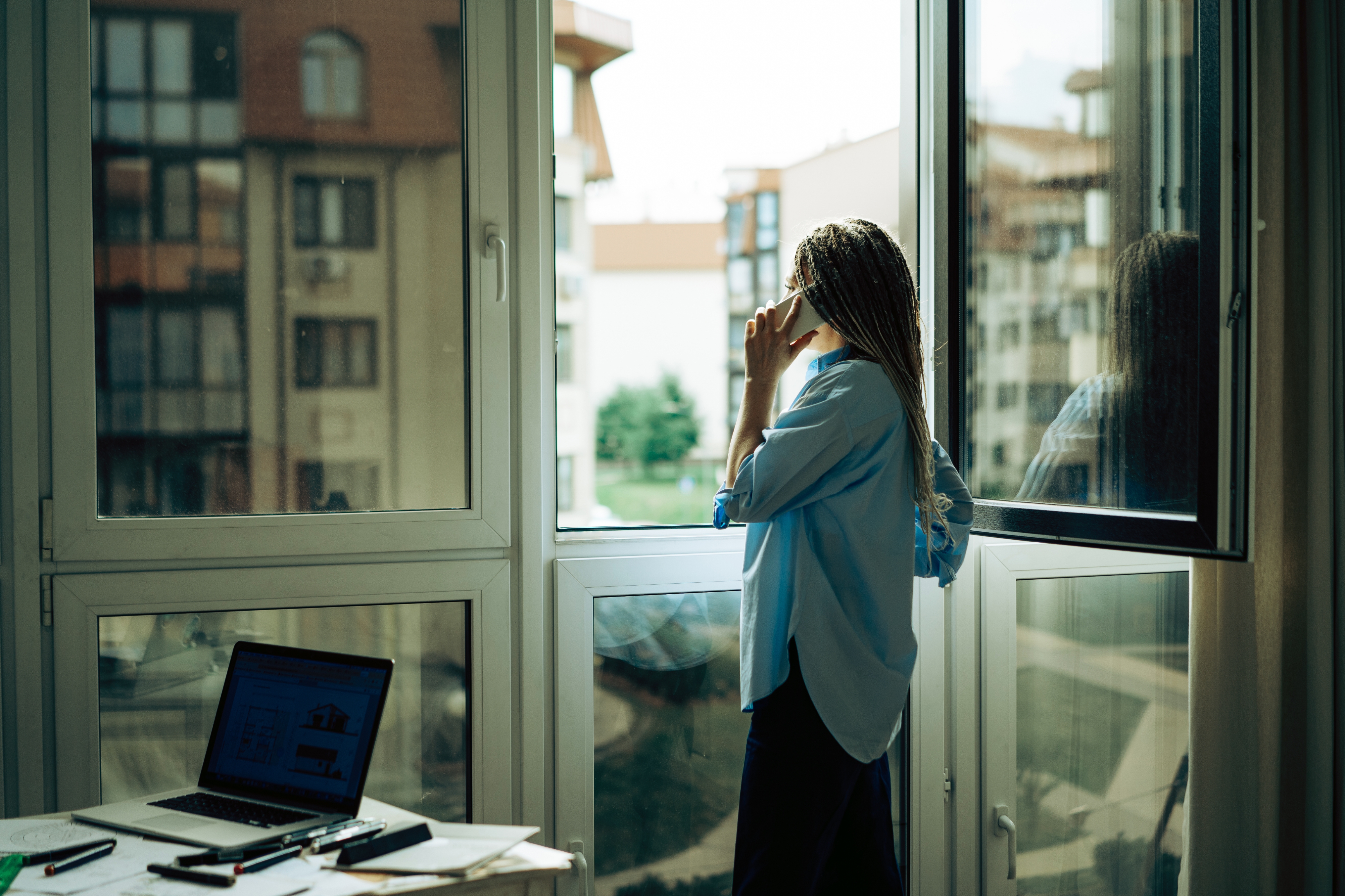 Person with long hair stands by a window, talking on the phone. A laptop and papers are visible on a nearby desk, suggesting a work-from-home setting