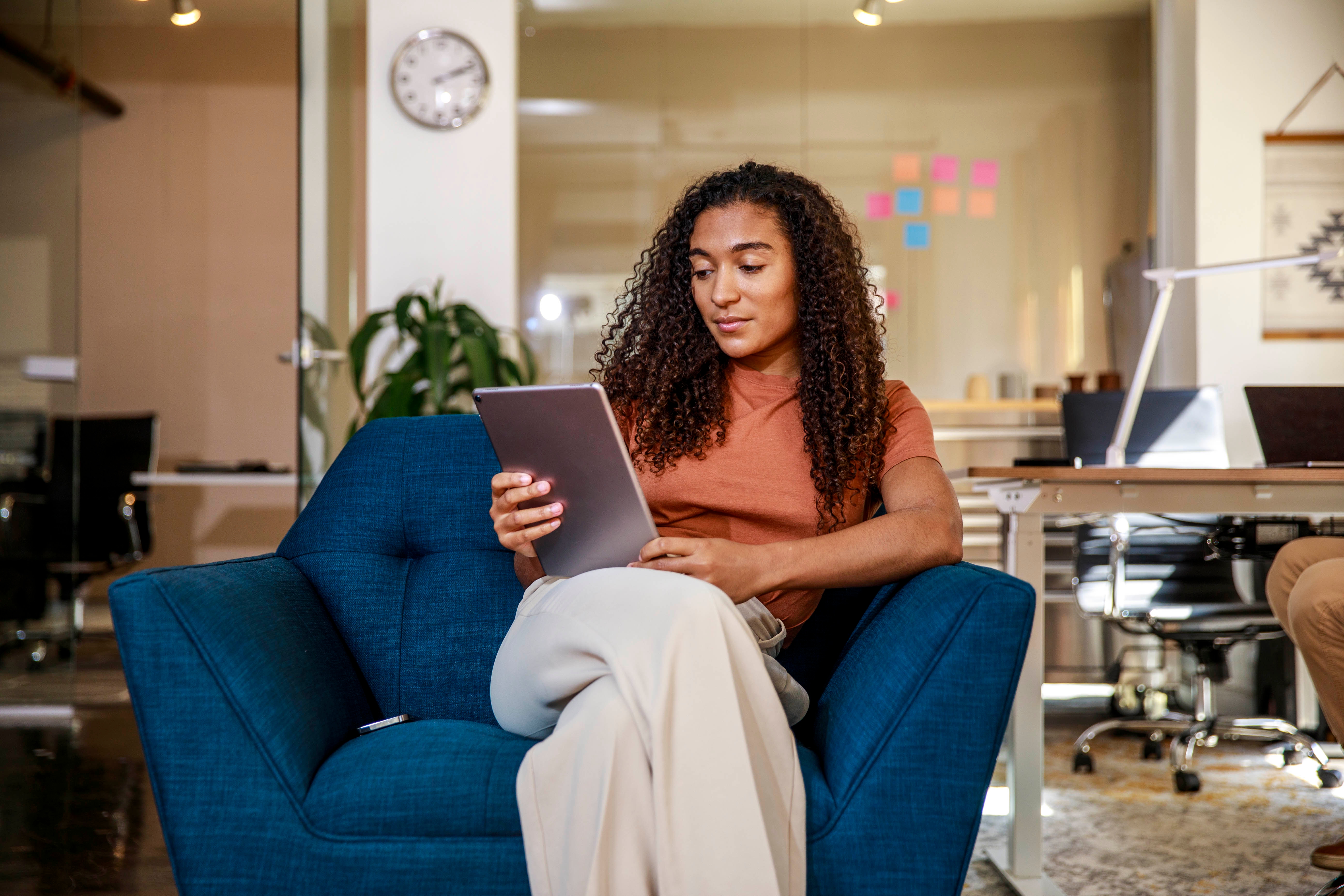 Person with curly hair sits on a blue chair, reading a tablet. Office background with desks and sticky notes on glass. Casual and focused setting