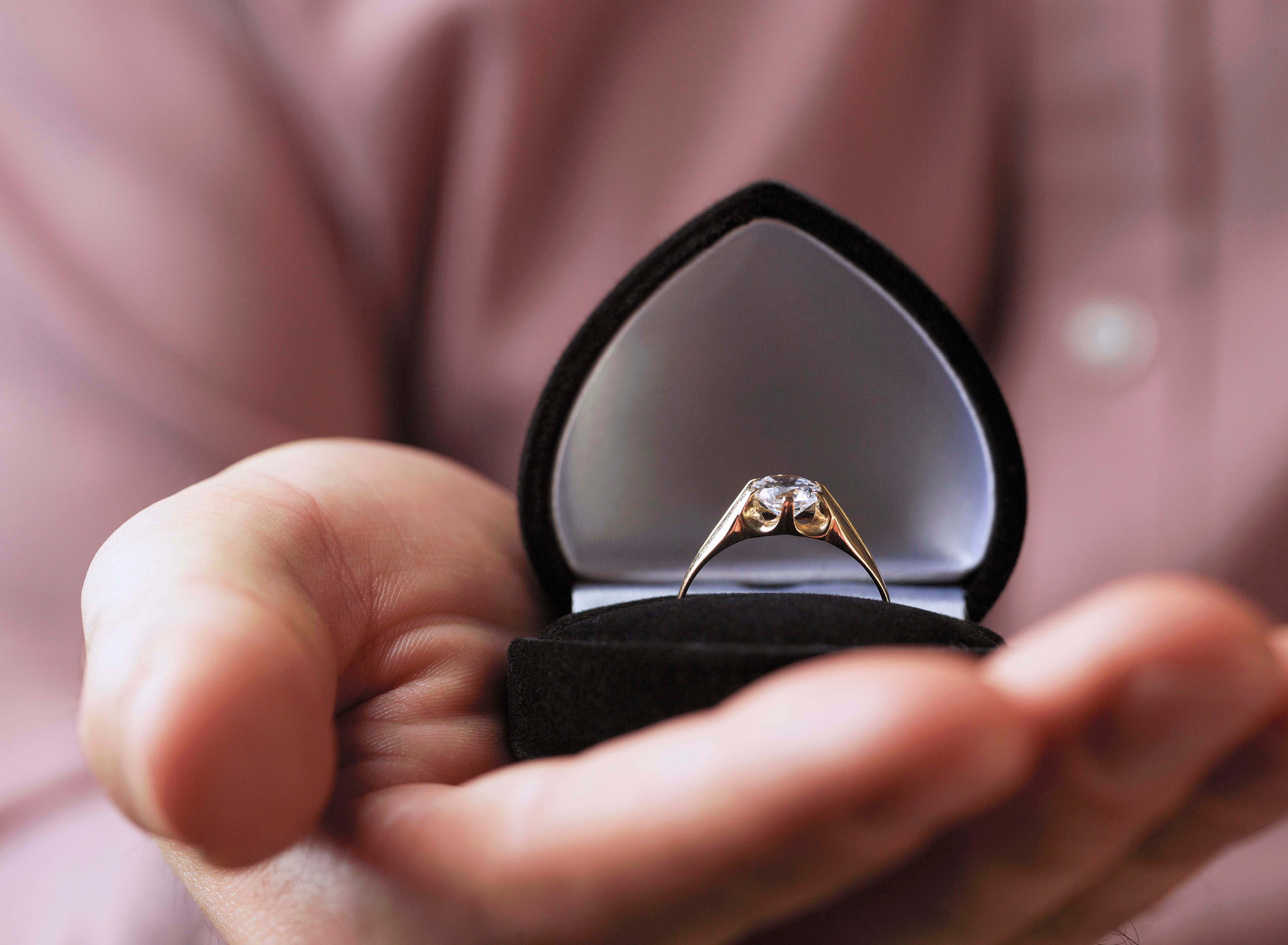 A hand holds an open ring box with a diamond ring inside, suggesting a romantic proposal or engagement theme