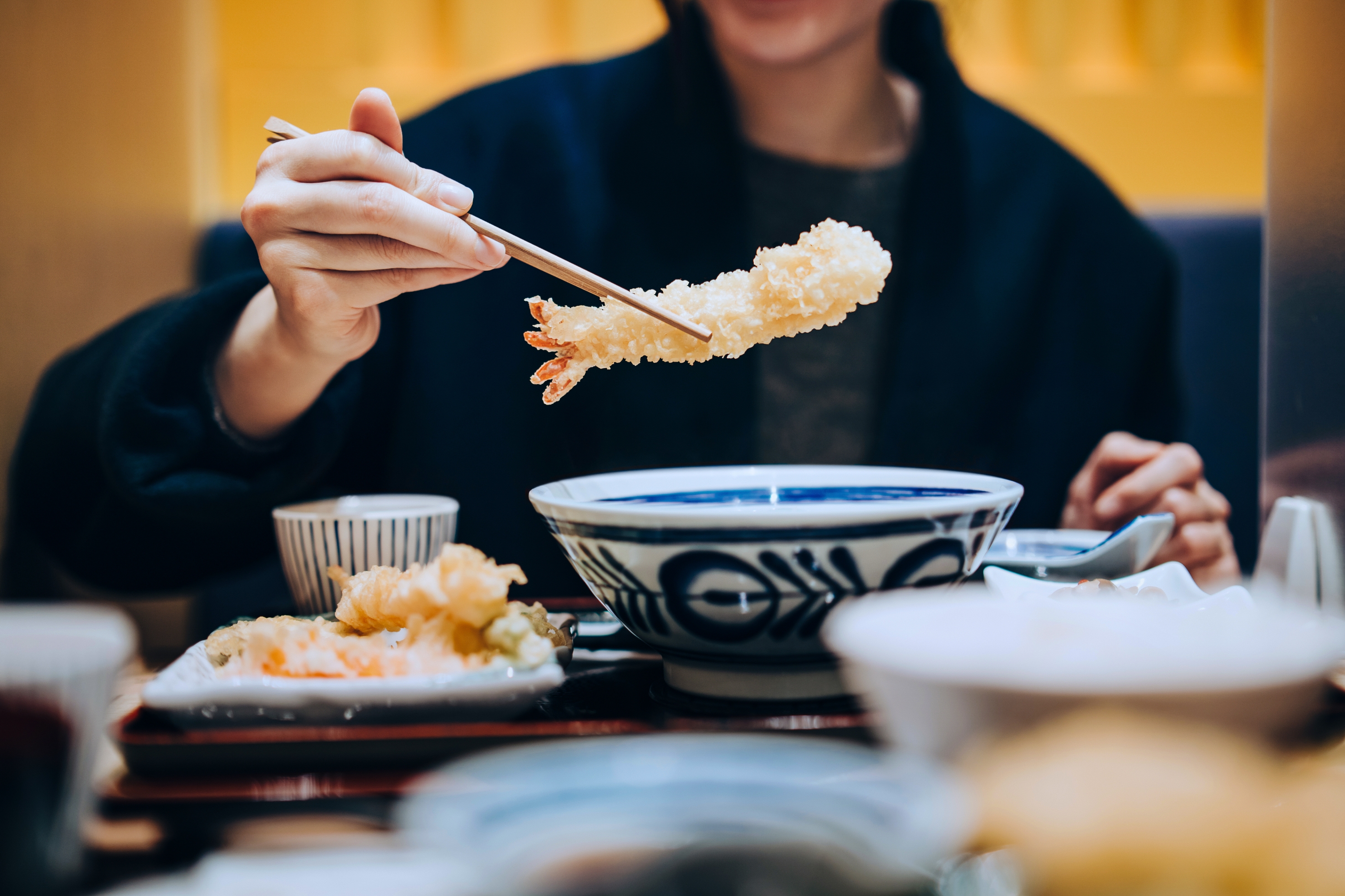 Person using chopsticks to pick up tempura shrimp over a bowl, suggesting a dining or romantic date setting