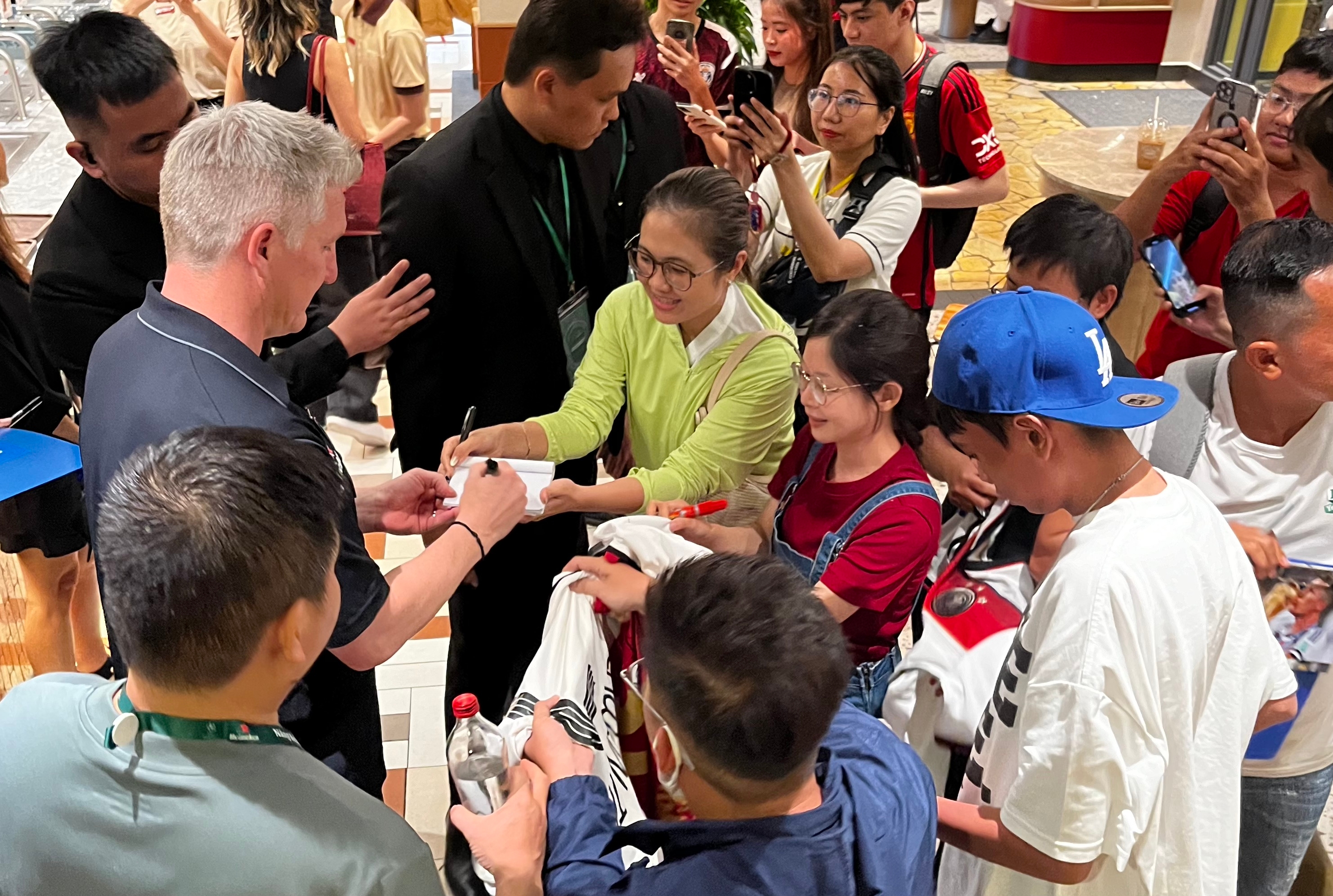 Person signing autographs for a gathered group of fans, with several holding jerseys and taking photos