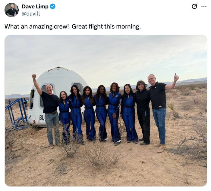 A group of people in blue jumpsuits and casual wear pose happily in front of a space capsule in a desert setting