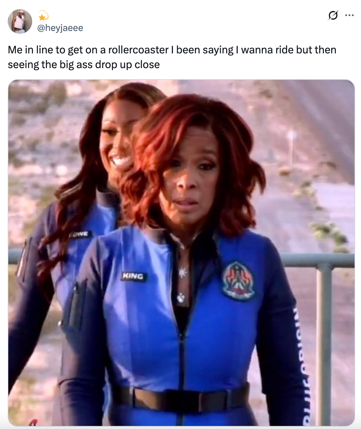 Two women in line for a rollercoaster, wearing blue jumpsuits, with one looking tense