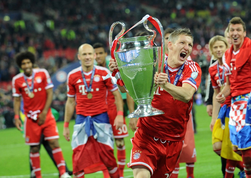 A soccer player joyfully lifts a trophy with teammates celebrating around him on a soccer field. Players wear team jerseys
