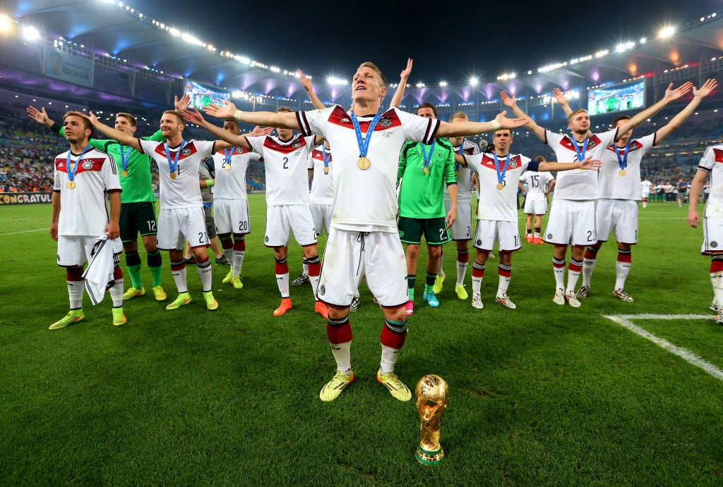 Soccer team celebrates on the field with arms raised. A player in front wears a gold medal, and the World Cup trophy is on the grass. Stadium lights shine