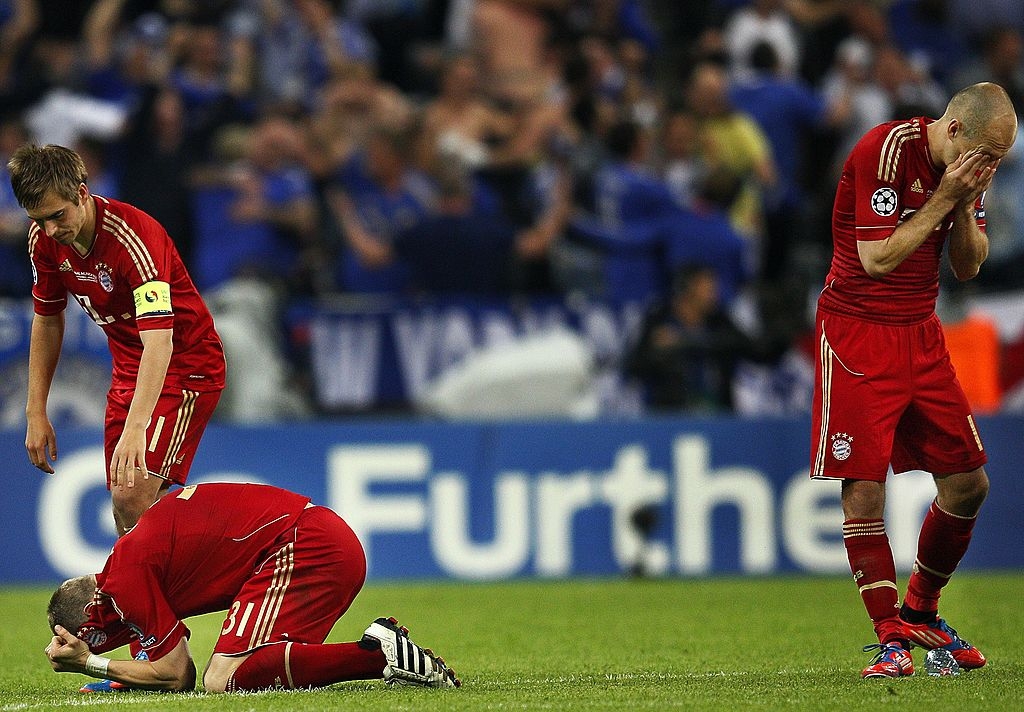 Three soccer players in red uniforms show disappointment on the field, one kneeling and covering his face