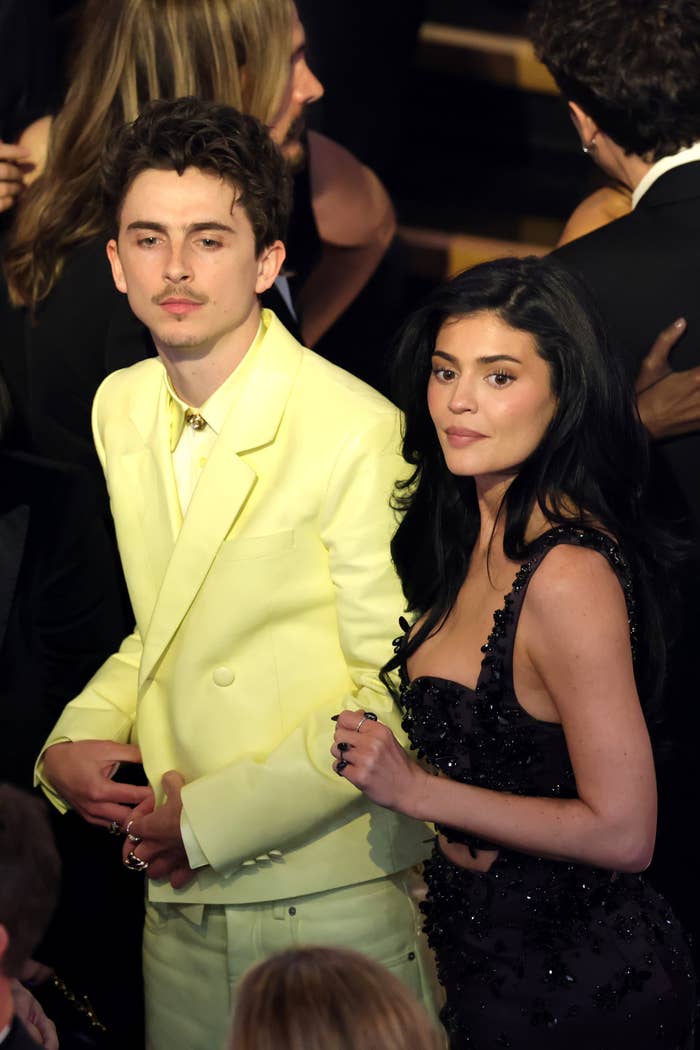 Two celebrities at an event: person on left in a light suit, person on right in a sparkling dress. They are in a crowded setting, facing the camera