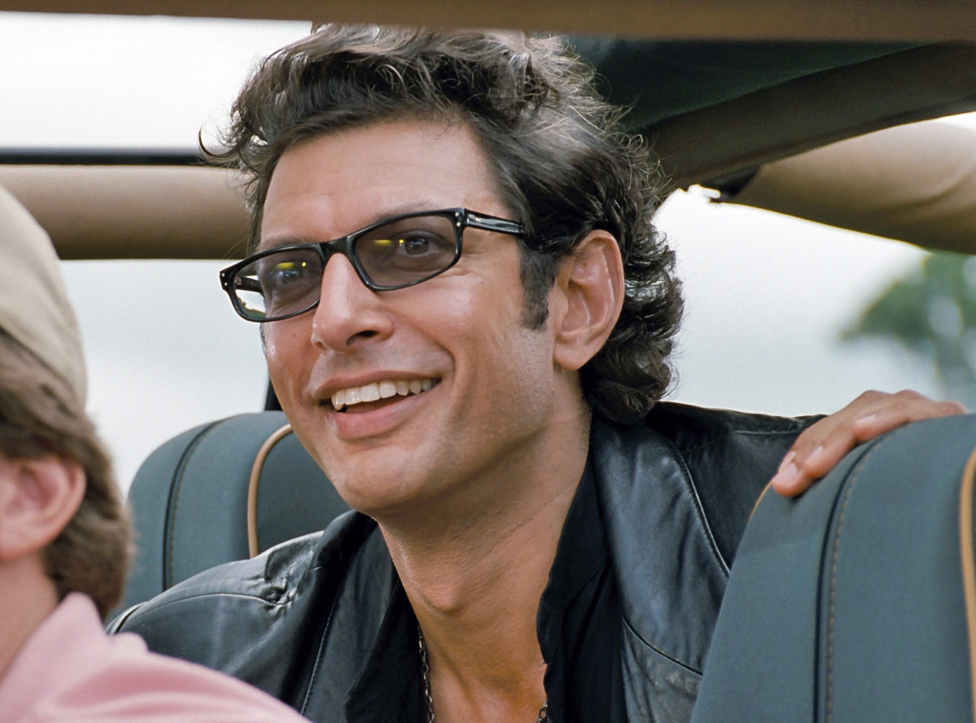 Person in a leather jacket and glasses smiles while seated in a vehicle, scene from a movie