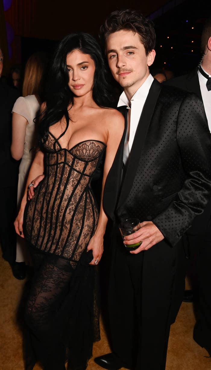Two individuals pose at a formal event. One wears a sheer, form-fitting dress; the other, a tailored suit. They stand close together, smiling slightly
